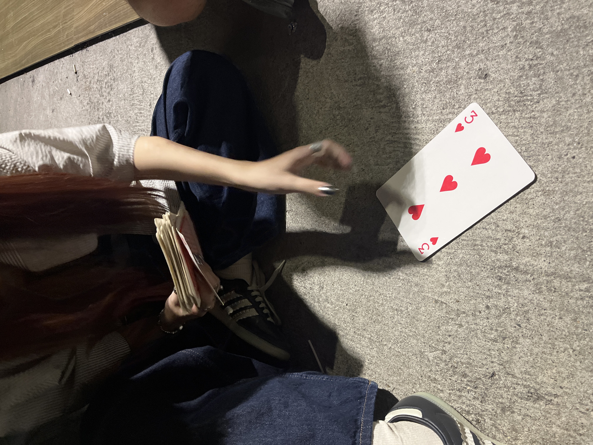 The image shows a close-up view of two people's legs and hands, sitting on a concrete surface. One person is wearing blue denim jeans and a white sock with a dark-soled shoe. The other person, whose legs are partially visible, is wearing dark blue pants. A hand with metallic silver nail polish is reaching towards a playing card on the ground. The playing card is the three of hearts, with three red heart symbols and the number "3" in the bottom right corner. The playing card is partially illuminated, casting a shadow. A stack of other playing cards is held by another hand in the upper portion of the image. The background consists of a rough concrete surface with some small debris and a wooden surface to the right. The lighting suggests it might be evening or nighttime, with artificial light sources. The setting appears to be an outdoor urban environment. The location is not identifiable as Vratsa, Bulgaria, or any other specific city.