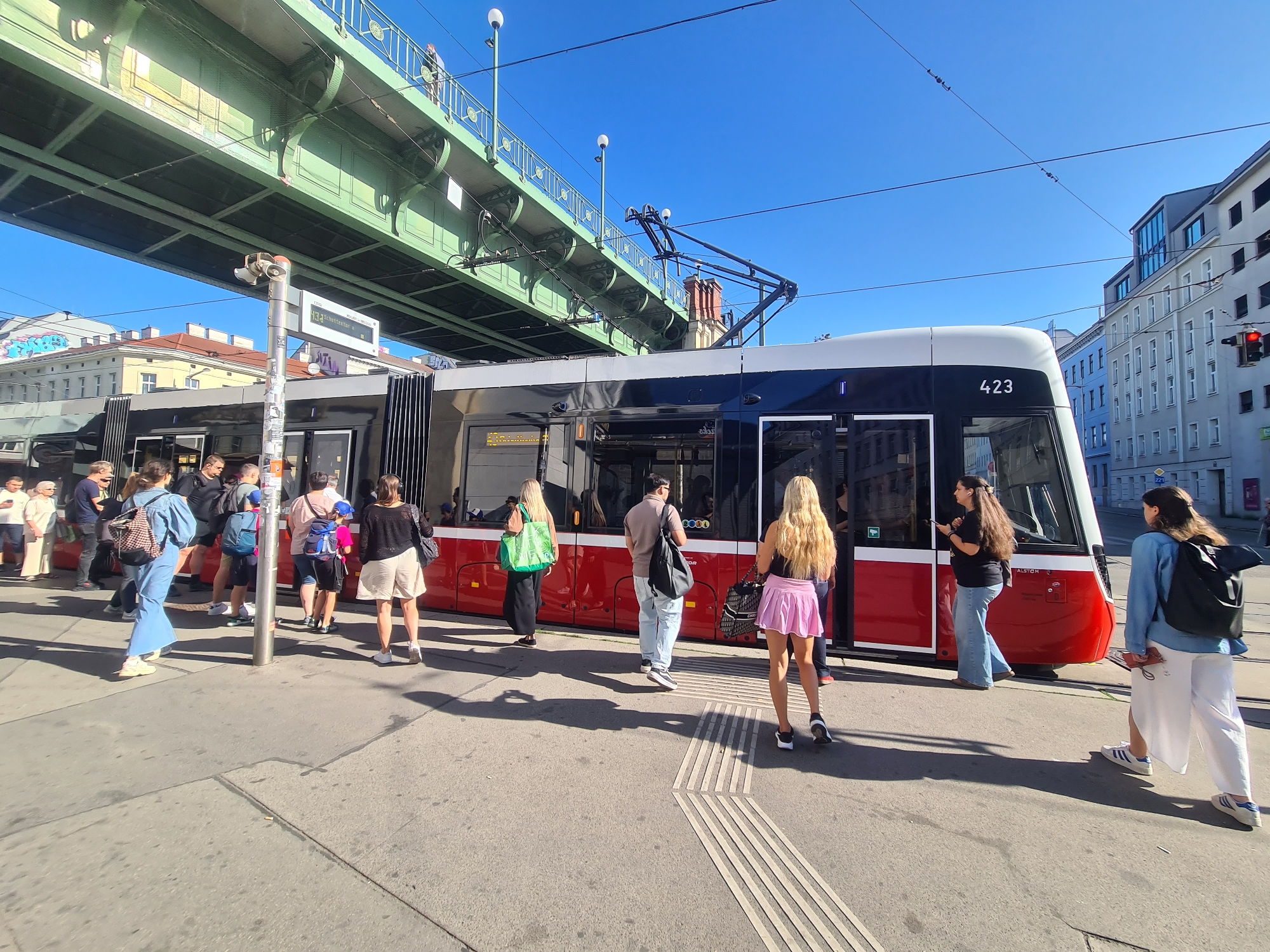 A tram with the number 423 is stopped at a station in Vienna, Austria, as a group of people wait to board or disembark. The tram is red and white, with large windows. Several people are standing on the platform, some with bags. A green elevated structure, likely a railway or public transport line, runs overhead. The sky is clear and blue, and the sun is shining, casting shadows on the ground. Buildings line the street in the background.