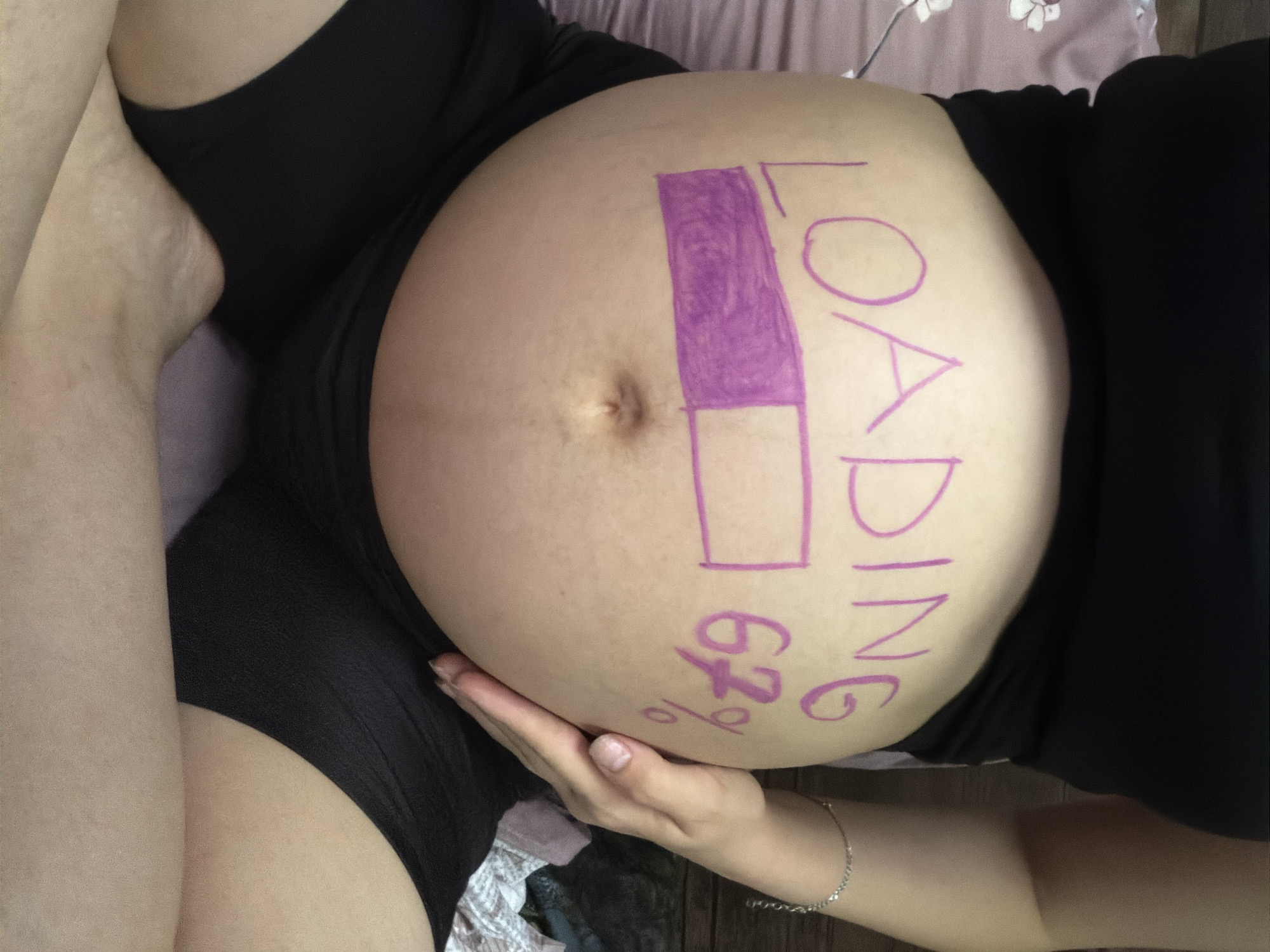 In a dimly lit indoor environment, likely a bedroom in Tbilisi, Georgia, a pregnant individual is seated cross-legged, presenting their abdomen for a photograph. The individual is wearing a black tank top and black shorts. Their pregnant belly is adorned with pink marker, depicting the word "LOADING" and a progress bar that is approximately 67% complete, indicated by the text "67%" to the right of the bar. The background consists of a patterned sheet and a wooden surface. The overall mood of the image is intimate and celebratory, suggesting anticipation for childbirth.