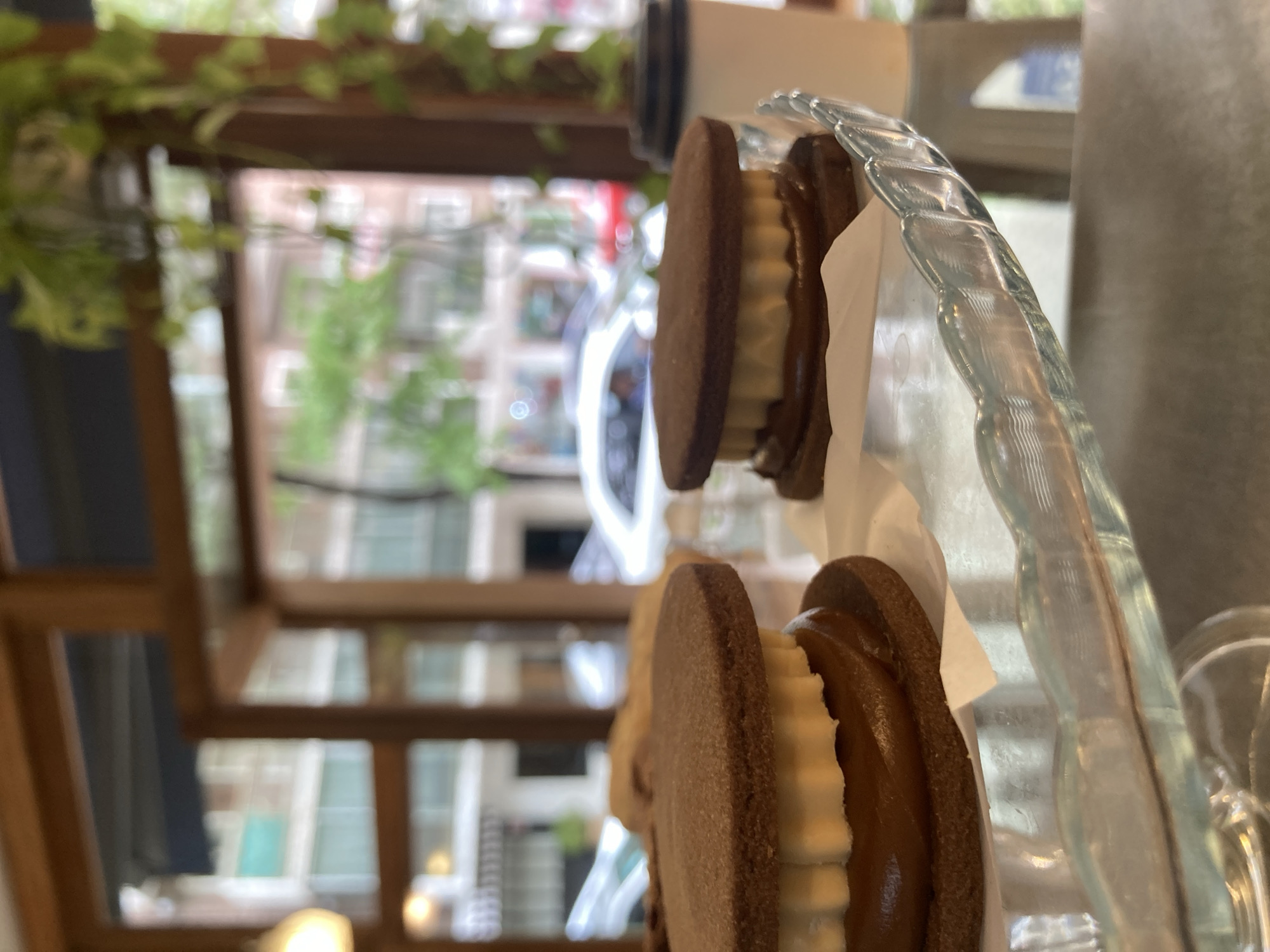 Captured at a cafe in an unspecified city, the image showcases two chocolate sandwich cookies. One cookie features a buttercream filling, while the other appears to be filled with caramel. They are elegantly presented on a glass dish, with a cup behind. Beyond the cookies, a window offers a glimpse of urban architecture. The scene is illuminated by natural light.