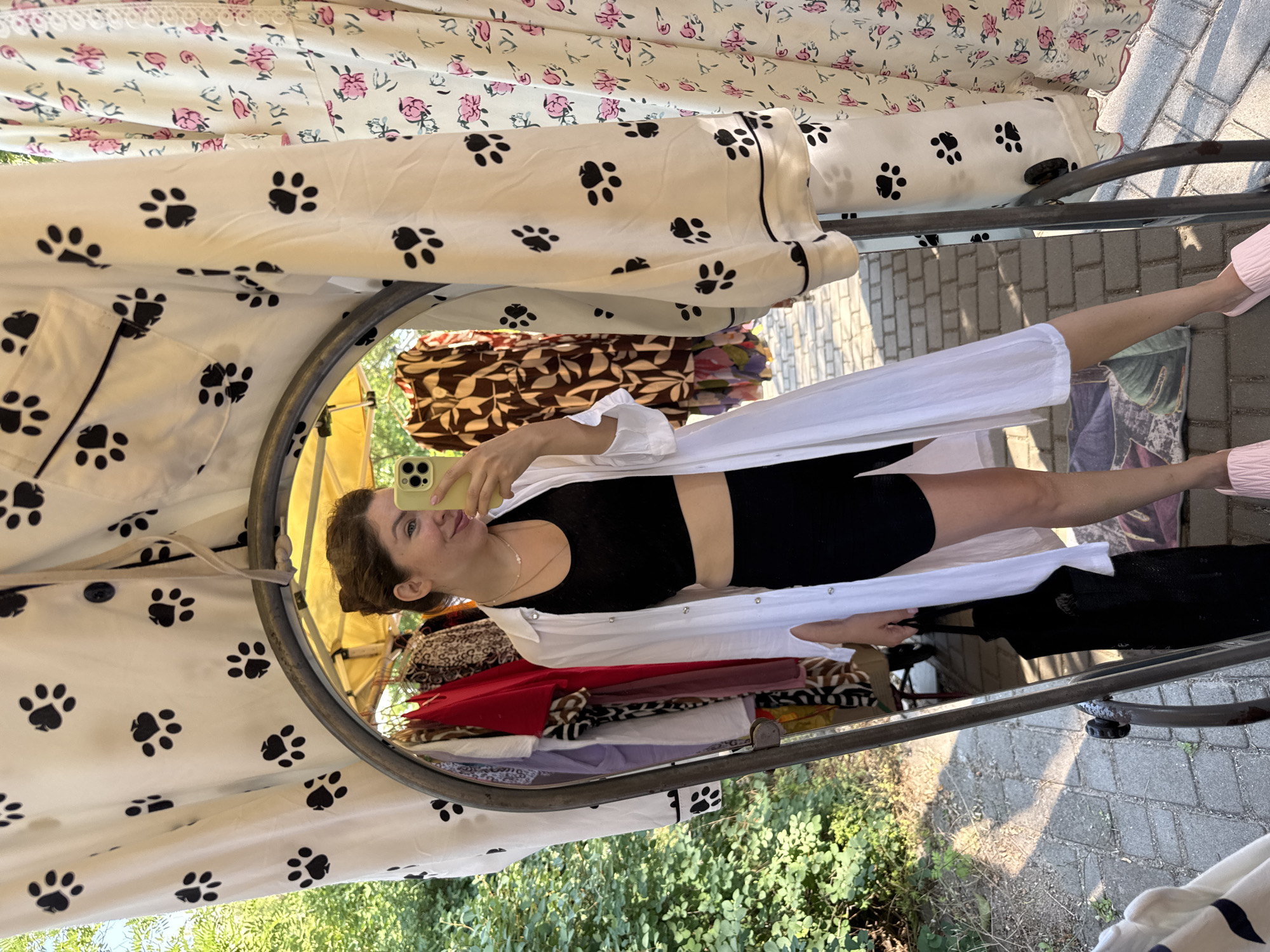A woman stands facing a mirror, taking a selfie with her smartphone. She is wearing a black crop top, black shorts, and a white unbuttoned shirt. She is also carrying a black tote bag in her left hand. The mirror reflects a clothing rack with various garments, and the background suggests an outdoor market setting with trees and paved ground, consistent with a location in Odesa, Ukraine. The woman has her hair tied up in a bun and is smiling at the camera. The clothing on the rack includes items with floral and paw print patterns.