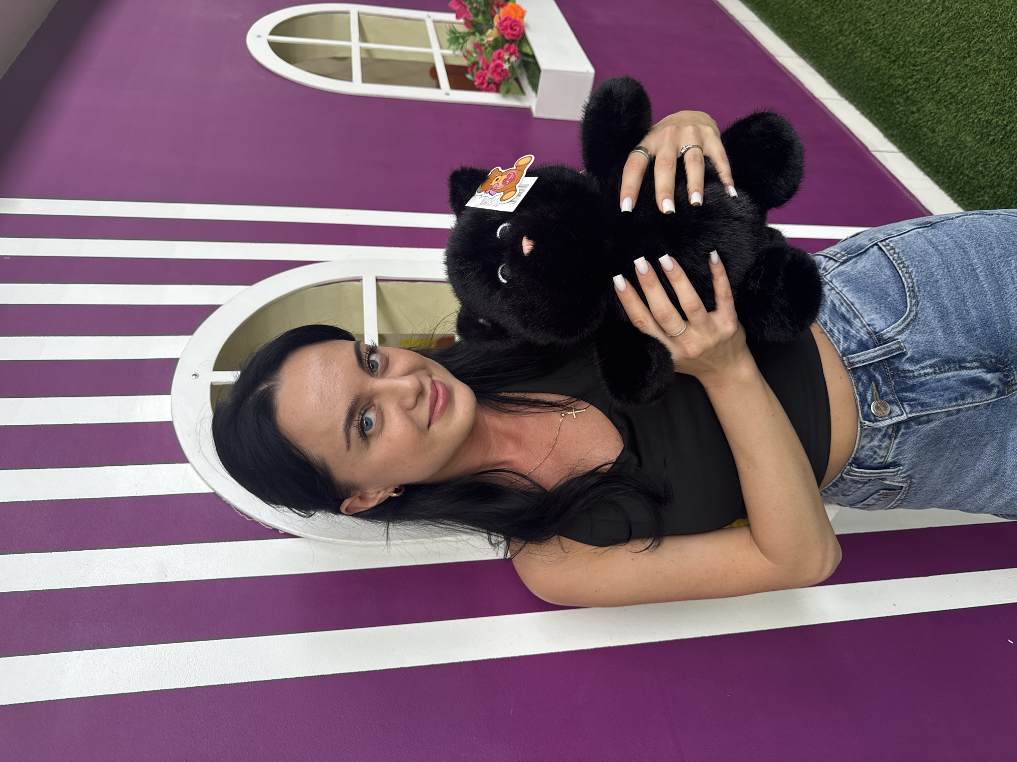 A woman lies on a purple and white striped surface, holding a black stuffed animal. She has long, dark hair and is wearing a black top and blue jeans. Her nails are painted white. The stuffed animal has a sticker on its head. In the background, there are white oval shapes that resemble windows, with some pink and orange flowers in one. Part of the surface appears to be covered in green astroturf. The lighting suggests it is daytime. The image appears to be taken indoors, possibly in a themed environment or play area. The woman's expression is relaxed and friendly. No visible text is present in the image.