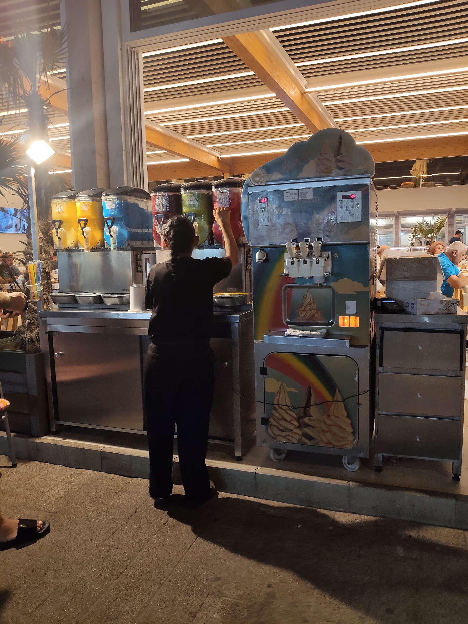 A person in dark clothing operates a soft-serve ice cream machine on a sidewalk in Constanța, Romania. The machine is adorned with a rainbow and ice cream cone graphics. Adjacent to it are several large dispensers holding colorful frozen beverages, including yellow, blue, red, and green. The scene appears to be outdoors, possibly at a kiosk or a restaurant patio, with tables and chairs visible in the background. The lighting suggests it is evening.
