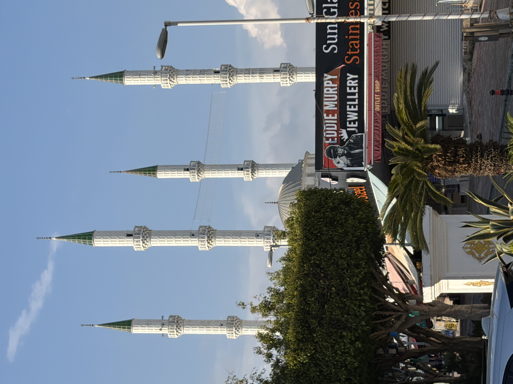 An outdoor, daytime scene in Alanya, Turkey, depicts a vibrant street with a prominent mosque and commercial establishments under a clear blue sky with scattered white clouds. The image is oriented vertically but should be viewed horizontally for correct perspective.
In the midground, four elegant white minarets with distinct green conical tops and gold finials rise against the sky. Their multiple balconies feature ornate detailing. Behind them, a mosque dome is partially visible. Lush green bushes and trees occupy the foreground and midground on the left, partially obscuring the lower sections of the minarets. Several tall palm trees are also visible, particularly on the right side.
On the right side of the street, various commercial buildings are present, featuring large, dark-colored signs. Visible text on these signs includes "Sun Gla" (likely "Sun Glasses"), "EDDIE MURPY" (likely "Eddie Murphy"), "JEWELLERY", and "Stainles" (likely "Stainless"). One sign for "EDDIE MURPY" also prominently displays an image of a person holding a camera. Below this, another sign reads "OUTLET SHOP". A tall street lamp extends into the sky from the right. A white car is partially visible on the far right. In the distance on the right sidewalk, a small person can be seen. The ground appears to be paved, suggesting a sidewalk or pedestrian area. The overall impression is that of a sunny day in a lively urban setting.