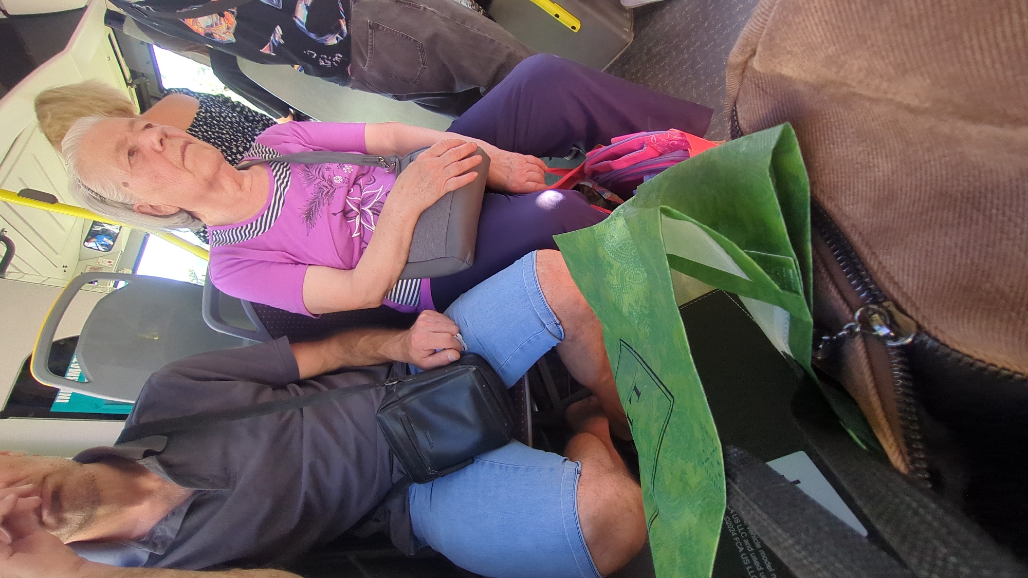 The image depicts an interior view of a public bus in Chișinău, Moldova. A man, partially visible from the chest up, is seated on the left side of the frame. He is wearing a dark grey polo shirt and light blue denim shorts. A black crossbody bag is slung over his shoulder. Beside him, an elderly woman is seated. She has grey hair and is wearing a purple t-shirt with a striped neckline and dark purple trousers. She has a grey shoulder bag and a pink bag is visible between her legs. In the foreground, a green reusable shopping bag is folded and placed on top of other bags. The bus interior shows grey seats and yellow safety bars. The lighting suggests it is daytime, possibly sunny.