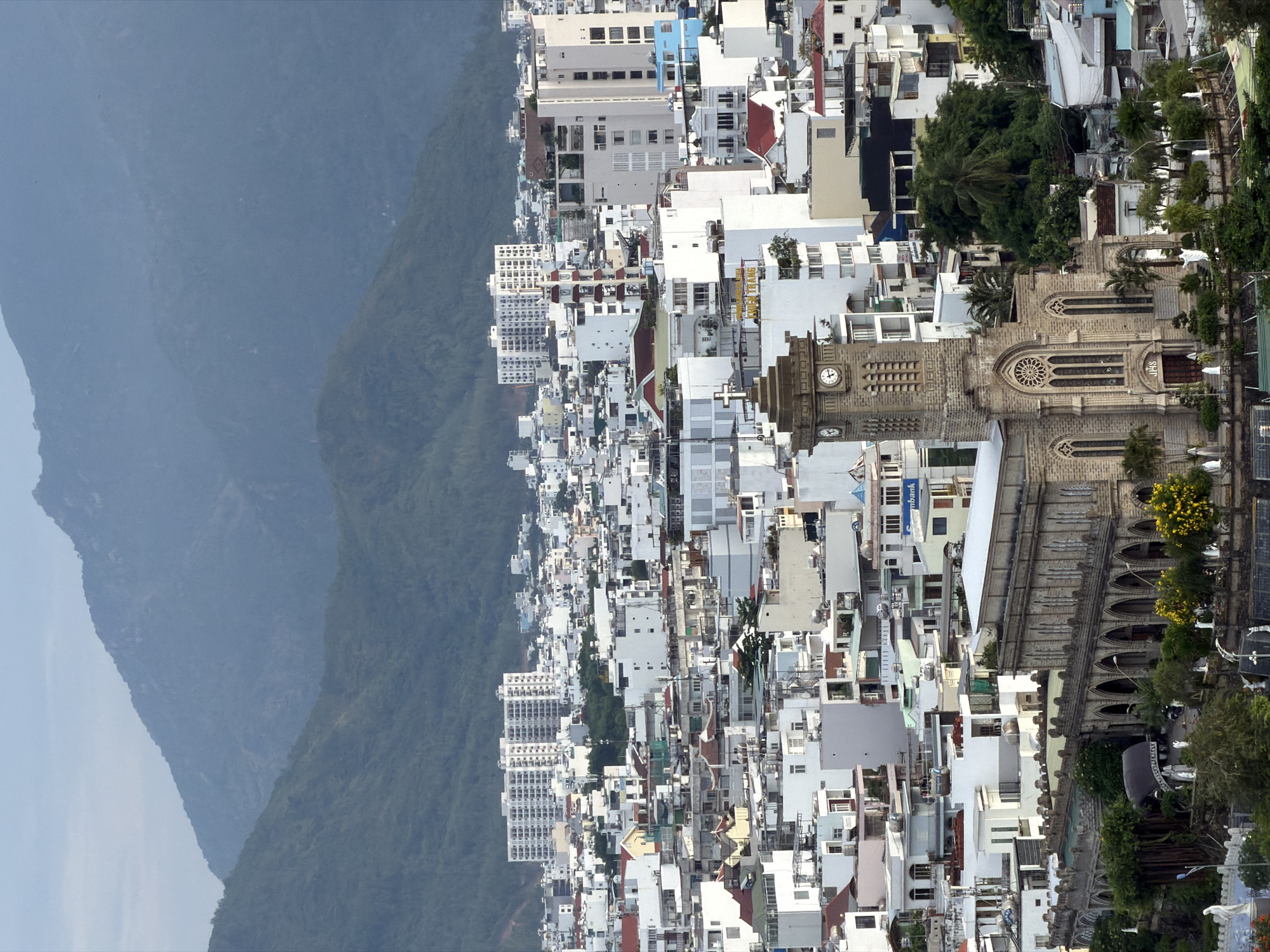 An elevated, panoramic view showcases a dense cityscape nestled between verdant mountains and a hazy sky, characteristic of Nha Trang, Vietnam. The scene is dominated by a multitude of buildings, primarily white or light-colored, featuring a mix of architectural styles, from multi-story residential blocks to modern commercial structures. Many rooftops are flat or reddish-brown, indicative of the tropical climate.
A large, imposing stone church stands prominently in the mid-ground, characterized by its Gothic revival architecture. It features a tall clock tower on the left, with the clock face displaying a time around 11:30 or 12:30. The church's facade includes intricate arched windows, a large rose window above the main entrance, and robust stone construction. Faint letters "DHS" are visible above the main entrance. Lush trees, including palm trees, are interspersed throughout the urban fabric, particularly around the church.
In the distance, towering mountains covered in dense green foliage rise majestically, partially obscured by a light atmospheric haze. Closer to the foreground, more clustered buildings and high-rises are visible. Visible text includes "VIET THANG" on a building sign and a partial "UBANK" on another. The lighting suggests a clear daytime, likely late morning or early afternoon, with soft, diffused light. The image captures a static urban landscape without any visible human activity.