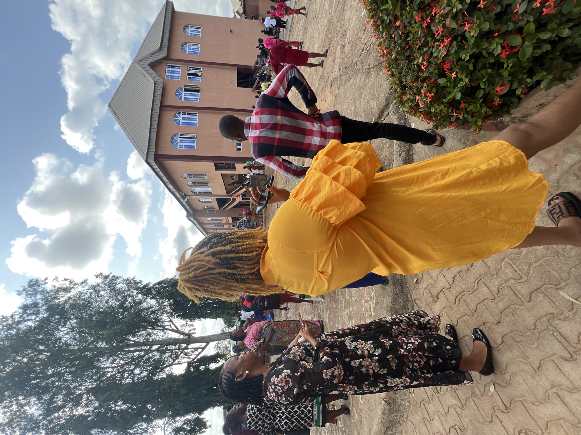The image depicts an outdoor gathering in front of a large, multi-story building, likely a church, in Nnewi, Nigeria. The building has a light brown facade, a grey pitched roof, and numerous arched windows with blue panes. A group of people are standing on a paved area in front of the building, some conversing and others looking around. In the foreground, a person wearing a bright yellow dress with ruffled sleeves is seen from the back, facing away from the camera. To the left, a woman in a black dress with a floral pattern is standing and looking to her left. To the right of the person in yellow, a man in a red, white, and blue checkered shirt and black pants is also seen from the back. Trees with green foliage are visible on the left side of the image, and a patch of flowering bushes with red blossoms is on the right. The sky is partly cloudy with a bright blue expanse. The overall scene suggests a community event or service taking place.