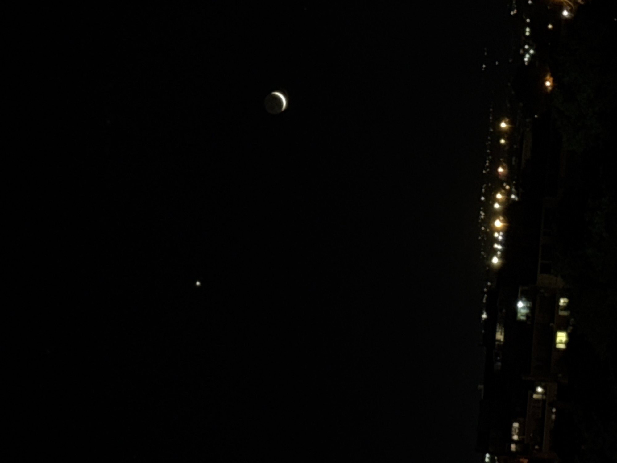 The image shows a night scene, likely in an urban environment. The moon, appearing as a crescent, is visible in the sky. Below the moon, a smaller, less distinct point of light is also apparent. To the right, blurred lights suggesting streetlights and building windows are seen, indicating a city or town. The image's vertical orientation and the presence of building lights suggest the photograph might have been taken from a higher vantage point. The location cannot be confirmed.
