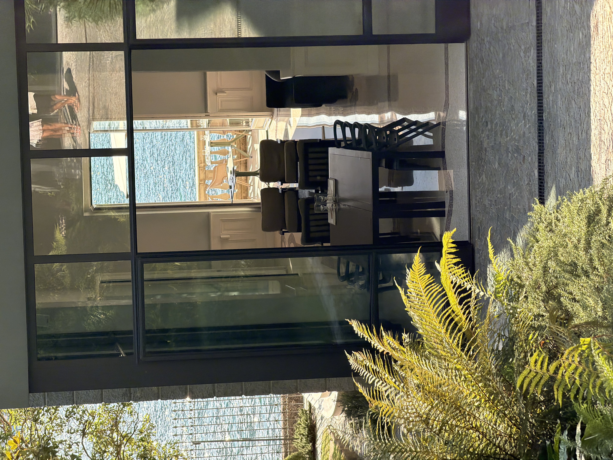 The image shows a view through a large glass window of a modern building located in Pognana Lario, Italy. In the foreground, lush green ferns are illuminated by sunlight. Beyond the ferns, a stone patio with a drain grate is visible. The interior of the building features a contemporary dining area with a dark rectangular table, several chairs, and a set of glasses. Two large armchairs are also present in the room. Through an open doorway, a balcony overlooks a body of water, likely Lake Como, with a table and chairs on the balcony. Two individuals, partially visible, are standing on a stone terrace in the distance, seen through the reflection in the glass. The architecture suggests a luxurious lakeside residence.