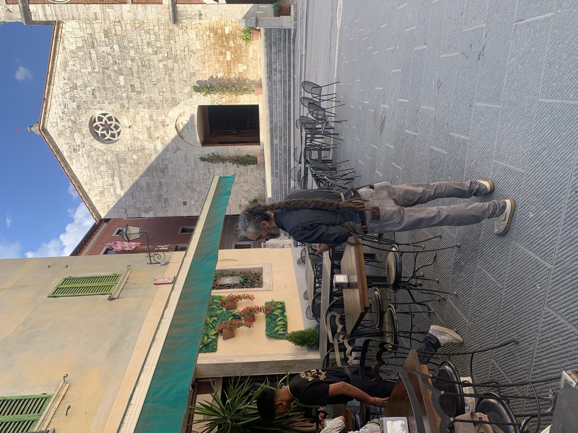 In San Quirico d'Orcia, Italy, a cobblestone plaza faces the facade of a stone church with a rose window and a cross atop its roof. Sunlight casts a distinct shadow from the church onto the ground. To the left, a yellow building houses an outdoor cafe with green awnings, tables, and chairs. A young man in a black t-shirt stands near the cafe's entrance, and an older man with a long braid down his back, wearing a dark shirt and grey pants, stands in the plaza, looking to his right.