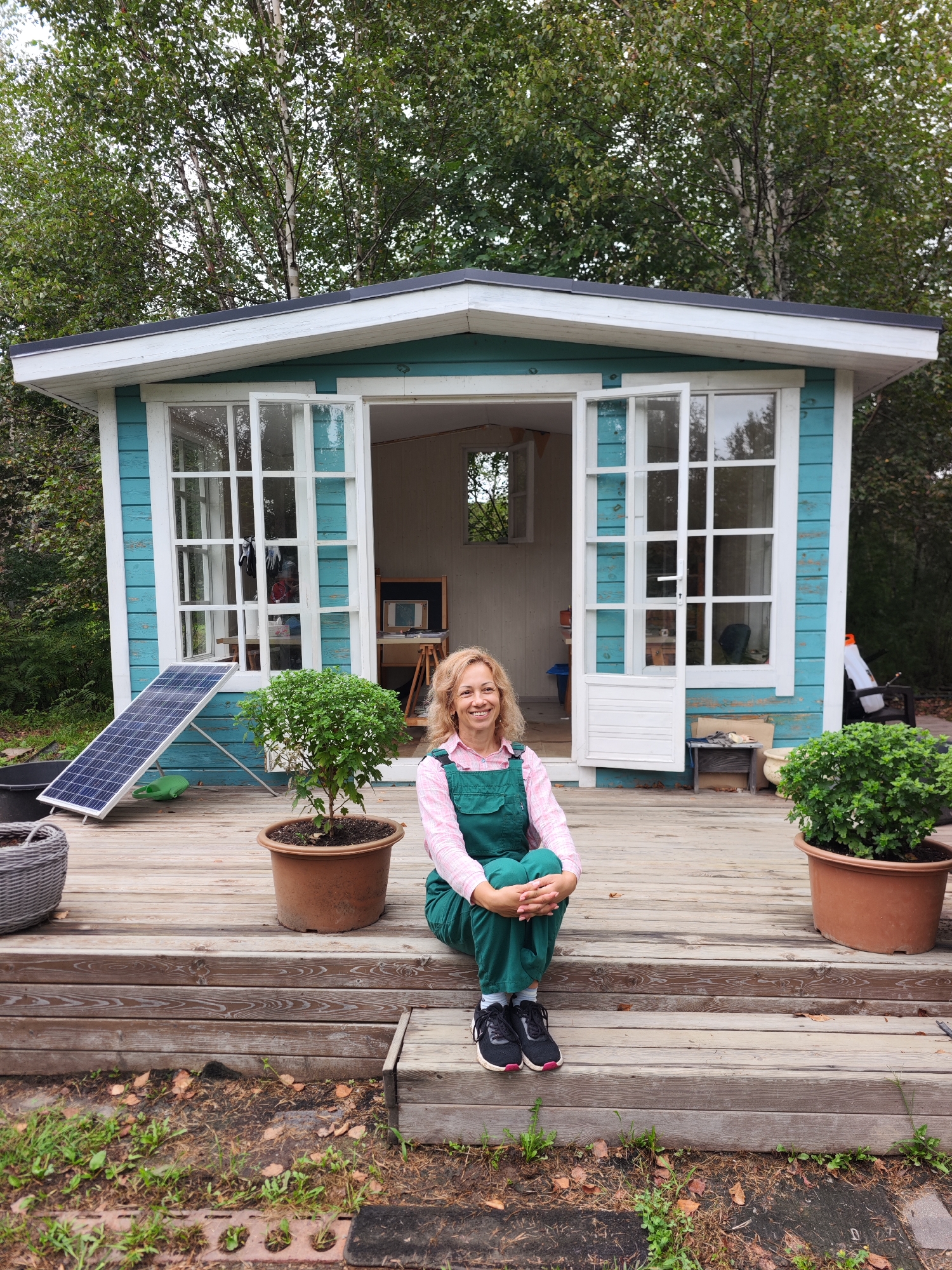A woman with blonde, wavy hair and a pink collared shirt under green overalls sits on the wooden steps of a small, teal-colored wooden shed with white trim. She is wearing black athletic shoes with pink accents. She is smiling and looking towards the camera. The shed has large French doors and windows that are open. Inside the shed, an easel with a canvas is visible. In front of the shed, to the woman's left, is a solar panel leaning against the shed and a small potted plant. To her right is a larger potted plant. The shed is situated on a wooden deck, and the surrounding area is an outdoor, possibly rural or garden setting with trees in the background and grass and fallen leaves on the ground. The overall scene appears to be in Solovej-Kljuch, Russia, given the context provided.