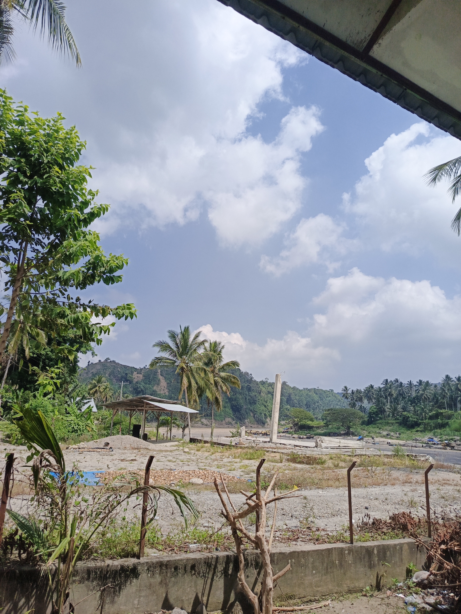 This bright, sunny image, taken in Simpangjaya, Indonesia, depicts a landscape that blends natural tropical vegetation with signs of human construction or development. The time of day appears to be mid-morning to early afternoon, given the clear blue sky dotted with scattered white cumulus clouds and the strong, even illumination.
In the immediate foreground, a low concrete wall with rusty metal fence posts runs horizontally. Dry brush and some green shoots grow along the wall and around the fence posts. A large, leafy green tree occupies the left side of the frame, with its canopy extending upward. A palm frond is visible in the top left corner, and a dark roof overhang is seen in the top right.
The mid-ground features a wide expanse of sandy and gravelly terrain, suggesting a dry riverbed or a construction site. Within this area, a modest, open-sided shelter with a light-colored corrugated roof stands on wooden posts. A large pile of sand or gravel is next to it. Further back, a tall, slender concrete pillar rises, appearing to be an unfinished element of a larger structure, possibly a bridge. Beyond this, a narrow body of water, likely a river, is visible, with what appear to be small vehicles or equipment on its banks.
The background is dominated by lush, green hills densely covered with tropical trees, including numerous palm trees. The scene offers a view of a serene yet active environment, potentially undergoing infrastructure development. There is no visible text in the image.