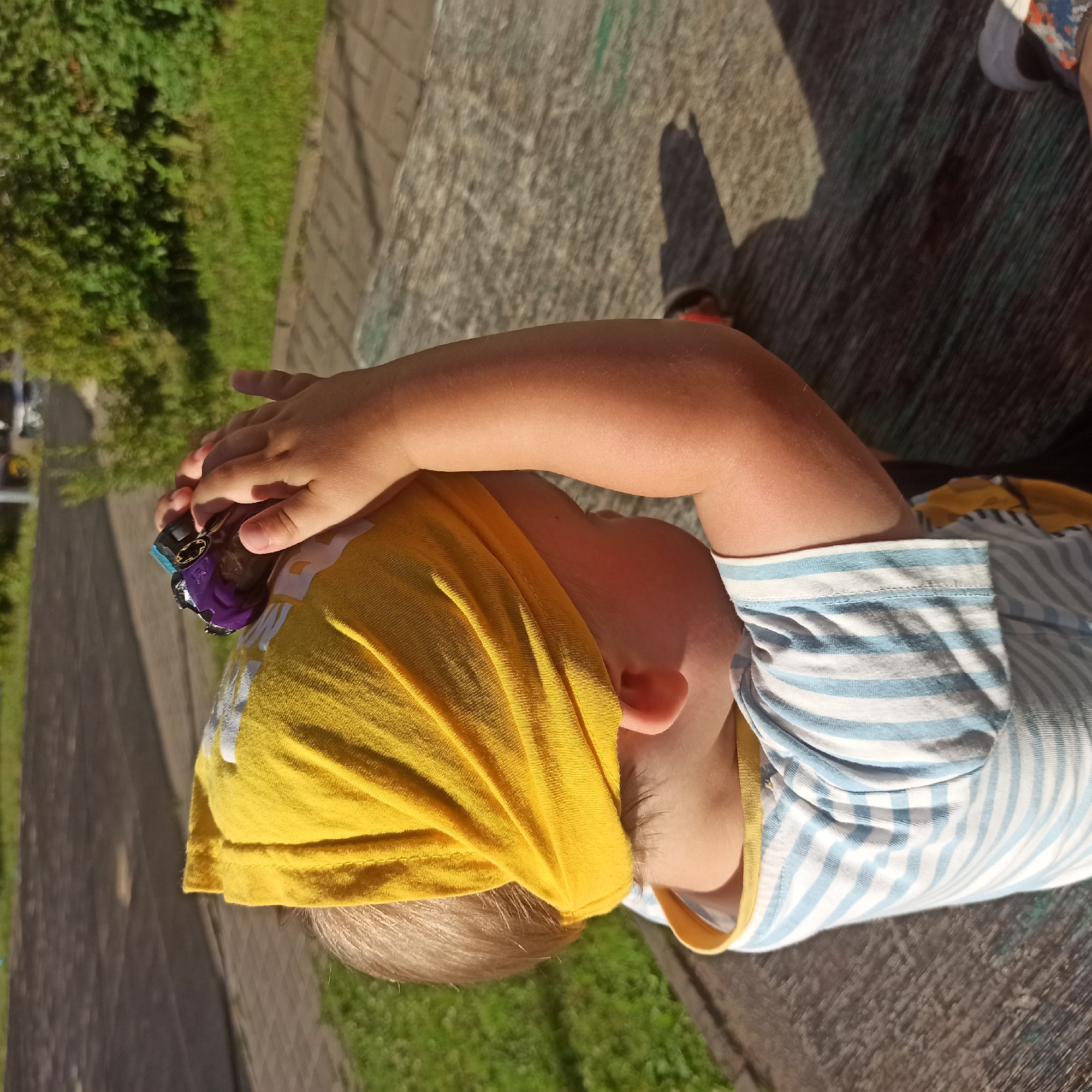 A young child wearing a yellow bandana and a blue and white striped shirt is sitting on a wooden surface outdoors. The child is holding a small purple and black toy car in their hands and appears to be looking at it intently. The background shows a paved area, grass, and some greenery, suggesting a park or outdoor recreational space. There are no discernible landmarks in the image that would indicate a specific location in Nizhny Novgorod, Russia.