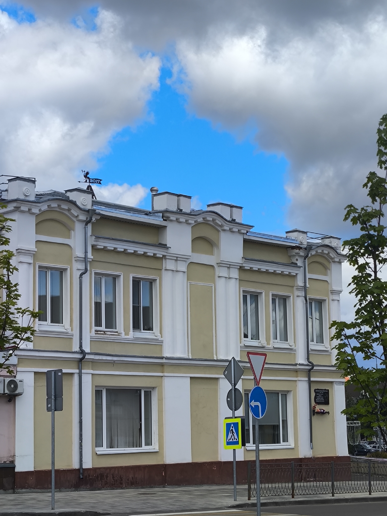 The image captures a segment of a pale yellow building with white decorative trim, located in Taldom, Russia. The building has a symmetrical facade with multiple windows, some of which are partially obscured by reflections. A weather vane shaped like a rooster with a flag displaying "2017" is visible on the rooftop. A series of traffic signs, including a yield sign, a turn arrow sign, and a pedestrian crossing sign, are positioned in front of the building. A small plaque is affixed to the wall of the building, accompanied by a floral arrangement. To the right of the building, a tree with green foliage partially obstructs the view. The sky above is a vivid blue, dotted with white and gray clouds. The foreground features a sidewalk, a street, and a decorative metal fence.