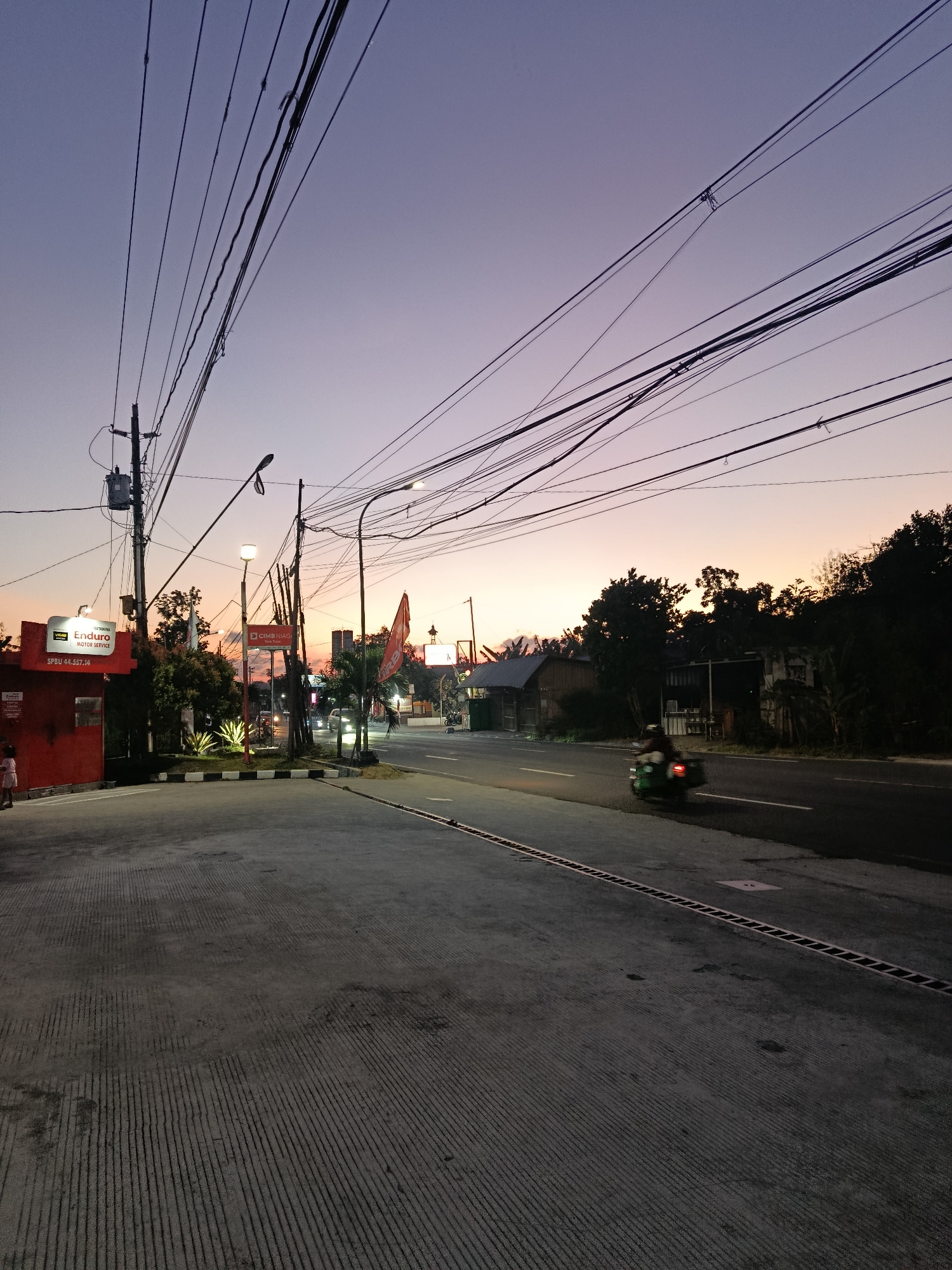 This is a ground-level, eye-level, medium shot photograph of a street scene at dusk in Sedayu, Indonesia. The sky is a gradient of deep purple near the top, transitioning to a soft orange and pink hue near the horizon. Numerous power lines crisscross the sky, casting a dense network of shadows. In the foreground, a paved area with textured concrete lines leads to a road. On the left side of the image, a red building with a sign that reads "PERTAMINA Enduro MOTOR SERVICE" is visible, along with a small child standing near its entrance. A utility pole with electrical equipment stands to the left of the building. The road in the midground is asphalt, with a white dividing line. A green scooter with a rider is moving away from the camera on the right side of the road. In the background, trees and various structures, including a building with a sign that appears to say "CIMB NIAGA," are silhouetted against the twilight sky. The overall atmosphere is that of a quiet, utilitarian urban fringe at the end of the day.