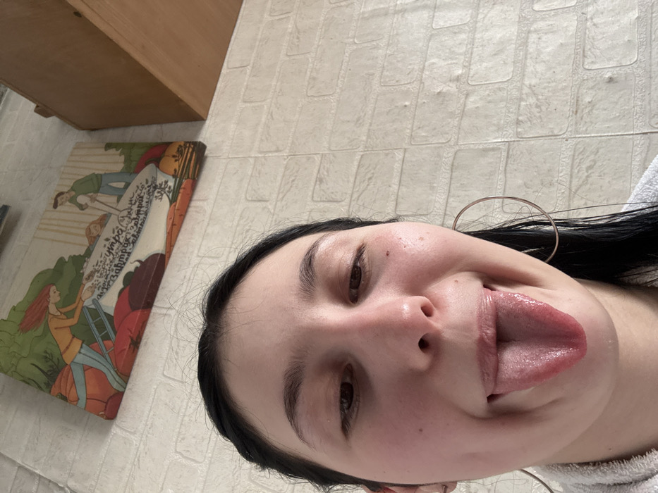 This image depicts an indoor residential setting, located in the city of Фоки, Russia. The primary subject is a young adult female, viewed from a close, upward-angled perspective, occupying the majority of the frame. She has dark hair, styled away from her face, and is looking downwards. Her tongue is extended from her open mouth in a playful or expressive gesture. Her cheeks and the bridge of her nose show some flushing. She wears large, thin hoop earrings in both ears. Her shoulders are covered by what appears to be a white, terrycloth material, possibly a towel or bathrobe.
The background features a light-colored, textured wall with a brick-like pattern. Mounted on this wall, above the female's head, is a horizontal rectangular piece of decorative wall art. This art illustrates a stylized scene with two figures: a woman with red hair on the left, seemingly interacting with a large, white trough or bowl that contains text, including the Russian word "завтрак" (breakfast). A man stands on the right side of the bowl. Below these figures, various fruits or vegetables (red on the left, orange on the right) are depicted. To the upper right of the wall art, a portion of a simple, light-colored wooden shelf is visible.