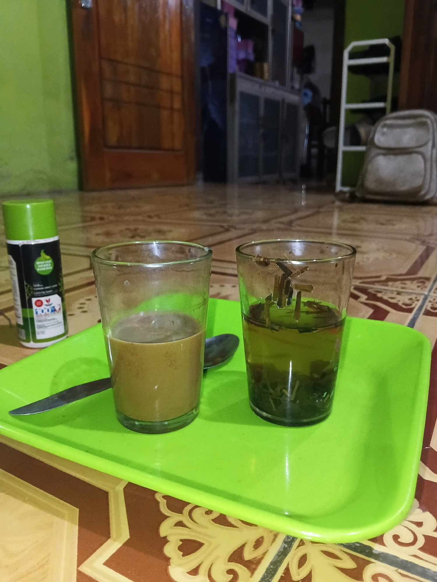The image shows a close-up view of two glasses of beverages on a bright green plastic tray. The glass on the left contains a light brown liquid, likely coffee, while the glass on the right contains a darker, greenish liquid with visible tea leaves, suggesting it is tea. A silver spoon rests on the tray between the two glasses. To the left of the tray, a green and white spray bottle with visible text is also present. The tray and its contents are positioned on a patterned floor, which appears to be tiled. In the background, blurred elements include a wooden door on the left, a set of cabinets and shelves with various items, and a white ladder-like shelf with a light-colored backpack on it to the right. The overall setting suggests an indoor environment, potentially a home in Margo Rukun, Indonesia.