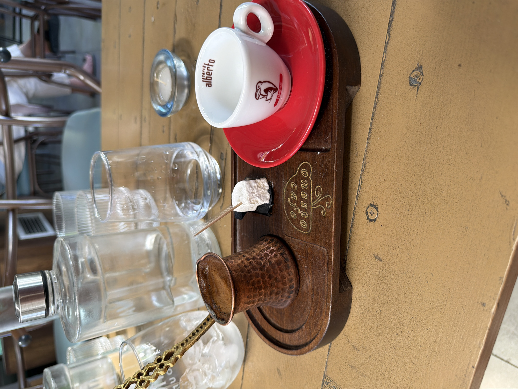 A close-up, high-angle shot shows a wooden tray with a small copper pot filled with coffee, a cube of Turkish delight, a white espresso cup on a red saucer, and a glass of water. A bottle of water, other glasses, and a small candle in a blue glass holder are also visible on a wooden table. The setting appears to be outdoors, possibly at an establishment named "onesto cafe" in Drama, Greece, based on the embossed text on the tray. The overall scene evokes a relaxed, cafe atmosphere.