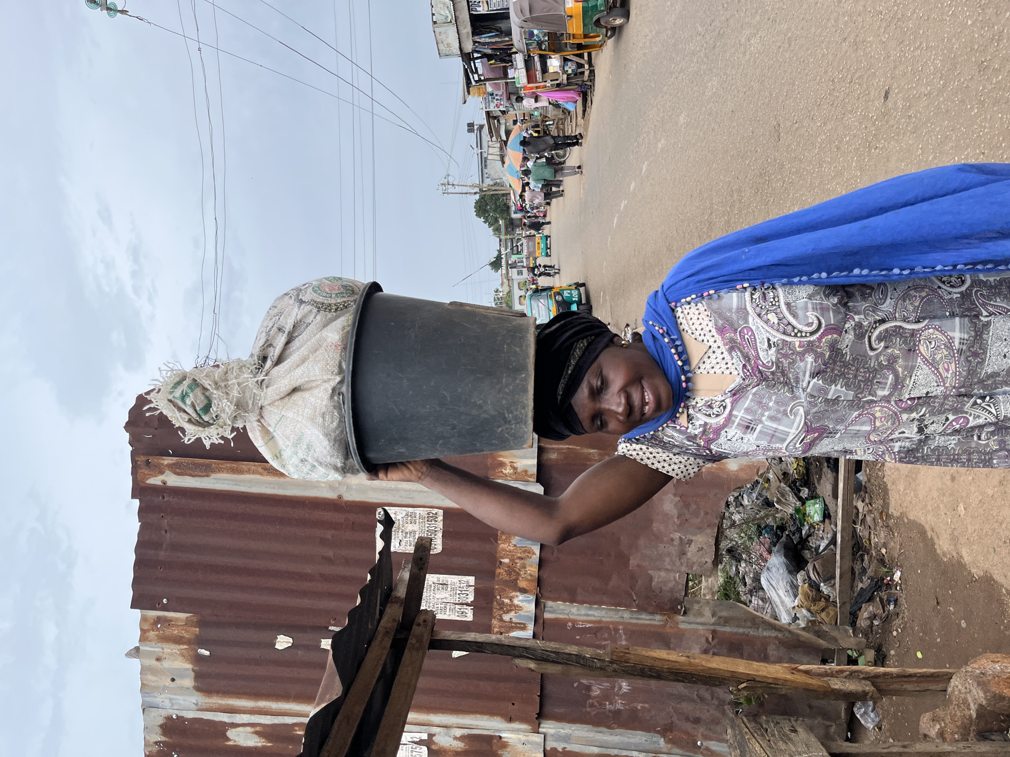 A woman stands near a corrugated metal fence with a black bucket and a sack on her head, on a street in Jos, Nigeria. In the background, a street scene unfolds with pedestrians, vehicles, and shops. The woman has dark skin and a blue scarf on her head and a patterned short-sleeved shirt. Behind her is a pile of garbage.
