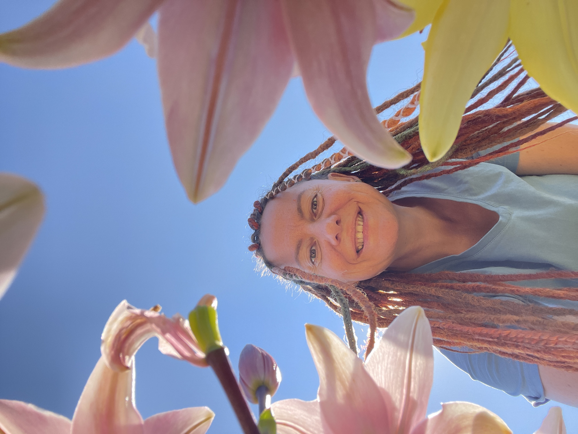 A low-angle shot captures a smiling woman with long, braided hair, possibly dreadlocks, in Tatarstan, Russia. She is looking up at the camera, framed by large, soft pink and yellow lily petals. The background is a clear blue sky. The woman is wearing a light blue t-shirt. The lighting suggests it is daytime, likely outdoors. The composition is dynamic, with the flowers creating a natural frame around the woman's face.