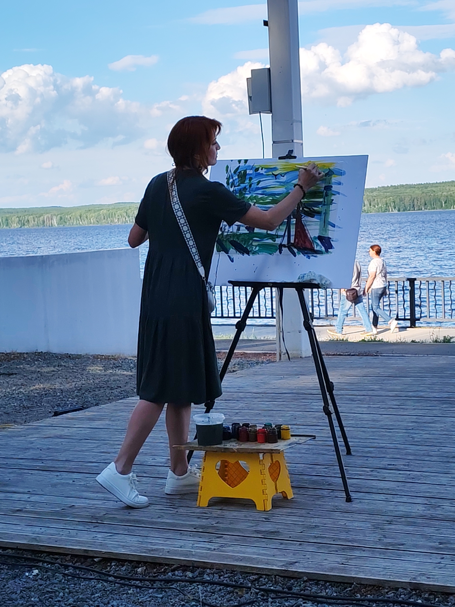 An artist with short, reddish-brown hair is painting en plein air by a body of water. She wears a dark green, knee-length dress and white sneakers. A white strap with a decorative pattern crosses her chest, and a small white bag is attached to it. She is actively painting on a canvas set on a tripod easel. The painting appears to be an abstract landscape with blue and green brushstrokes.
Beside the easel, a bright yellow step stool holds an assortment of paint containers and a brush. The artist's right hand holds a paintbrush, and her left hand is near the canvas.
In the background, a wide expanse of blue water stretches towards a treeline on the horizon. A white railing separates the wooden boardwalk from the water. Further in the background, another person, a woman wearing jeans and a white shirt, walks along the boardwalk.
The scene is set on a wooden boardwalk. The sky is bright blue with fluffy white clouds, suggesting a pleasant day. The lighting indicates it is daytime, likely afternoon. The presence of the water, trees, and boardwalk suggests an outdoor recreational area or a scenic overlook. The artist's focused posture and the act of painting convey a sense of creativity and engagement with the environment. There is no visible text in the image.