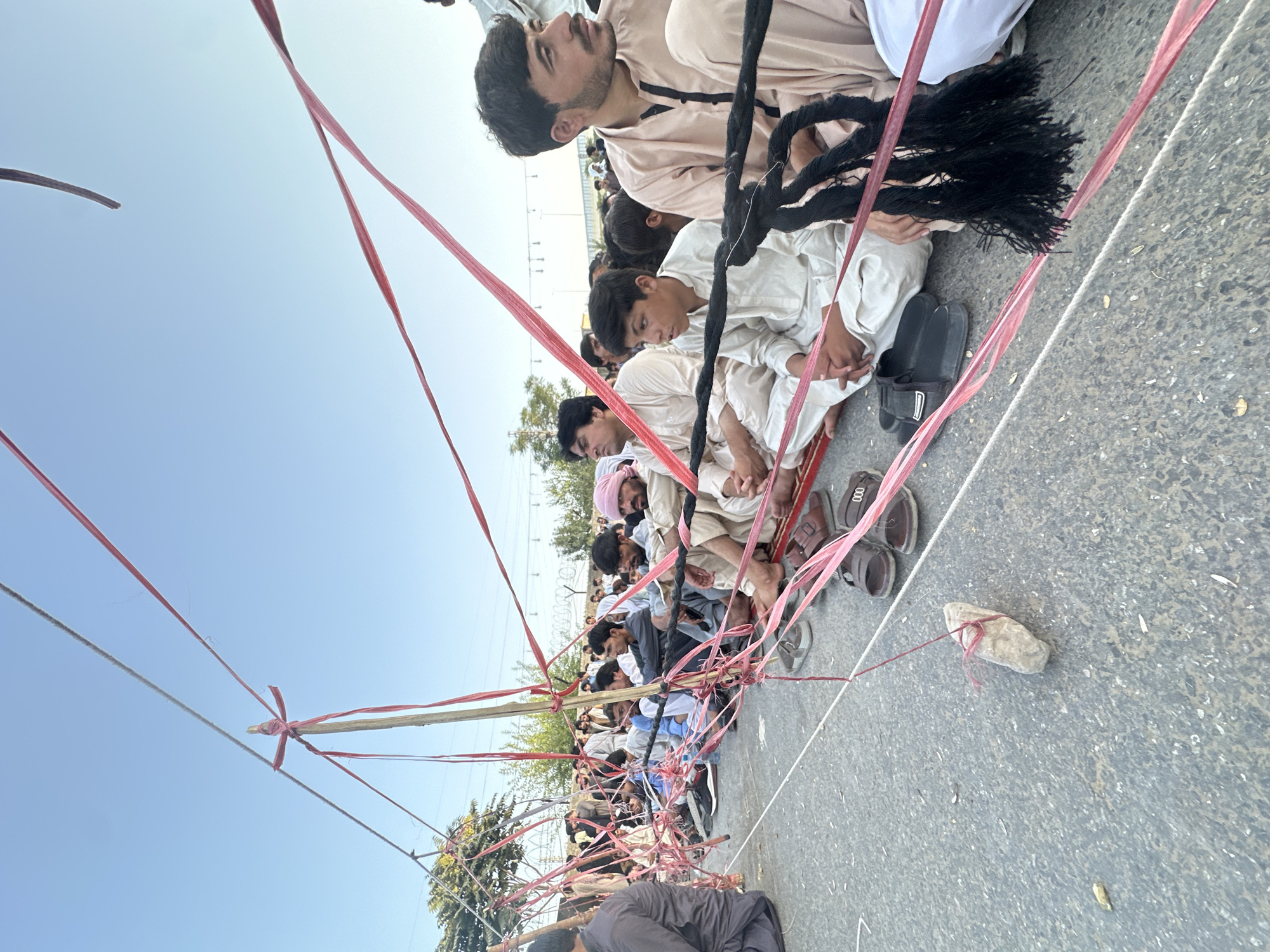 In Quetta City Tehsil, Pakistan, a group of men are seated on a paved surface, which appears to be a road or an outdoor gathering area. The men are dressed in traditional Pakistani attire, primarily shalwar kameez, with some wearing turbans. They are sitting in rows, with a noticeable barrier constructed from wooden poles and taut red ribbons strung between them. A thick, black rope is also integrated into this barrier in some sections. Several pairs of sandals and shoes are visible on the ground in front of the seated men, suggesting they have removed them. In the foreground, a stone is tied with red ribbon and suspended from one of the strings, possibly serving as an anchor or a marker. The background shows trees, a chain-link fence, and some distant buildings or infrastructure. The sky above is clear and blue, indicating daylight. The overall scene depicts an organized gathering or protest.