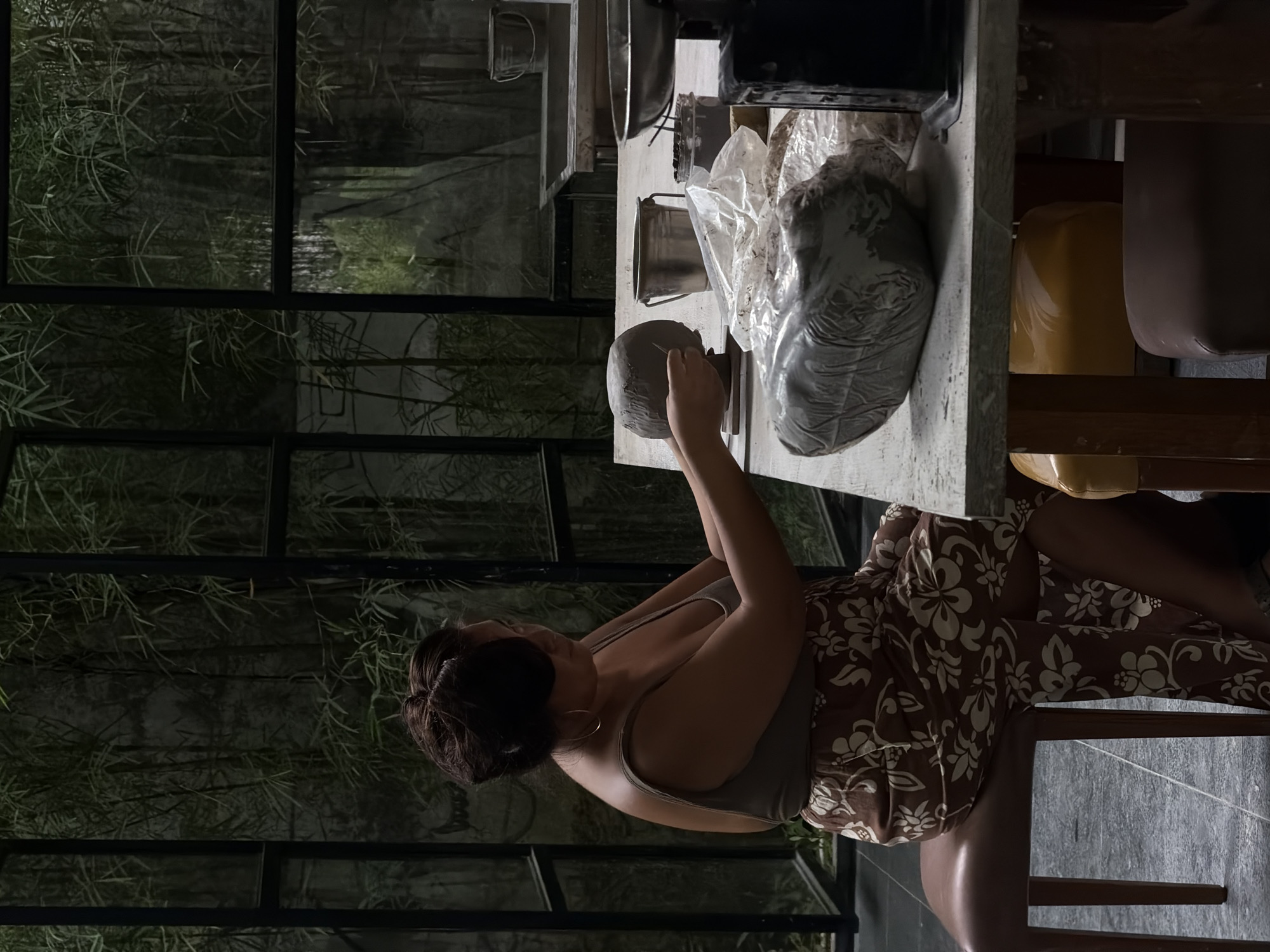 An indoor scene in Singakerta, Indonesia, features an adult female seated at a workstation, engaged in sculpting. The woman, positioned mid-frame and facing right, wears an olive-green sleeveless top and a brown long skirt with a white floral pattern. She has dark hair styled up and wears gold hoop earrings. Her hands are actively shaping a rounded gray clay form with a sculpting tool on a light-colored wooden table.
On the table, alongside the primary clay form, are several plastic-wrapped bags of gray clay, a small silver metal bucket, a glass jar containing a dark substance, and a metal tray. A dark-colored piece of equipment, possibly a scale, is partially visible on the far right of the table. The woman is seated on a brown cushioned stool. Under the table, parts of a yellow cushioned stool and another dark cushioned stool are visible.
The background consists of large, dark-framed glass windows that reveal a dense outdoor environment dominated by green bamboo plants. Behind the table, further interior elements include stainless steel sinks or work surfaces. The floor throughout the visible area is dark gray tiled.