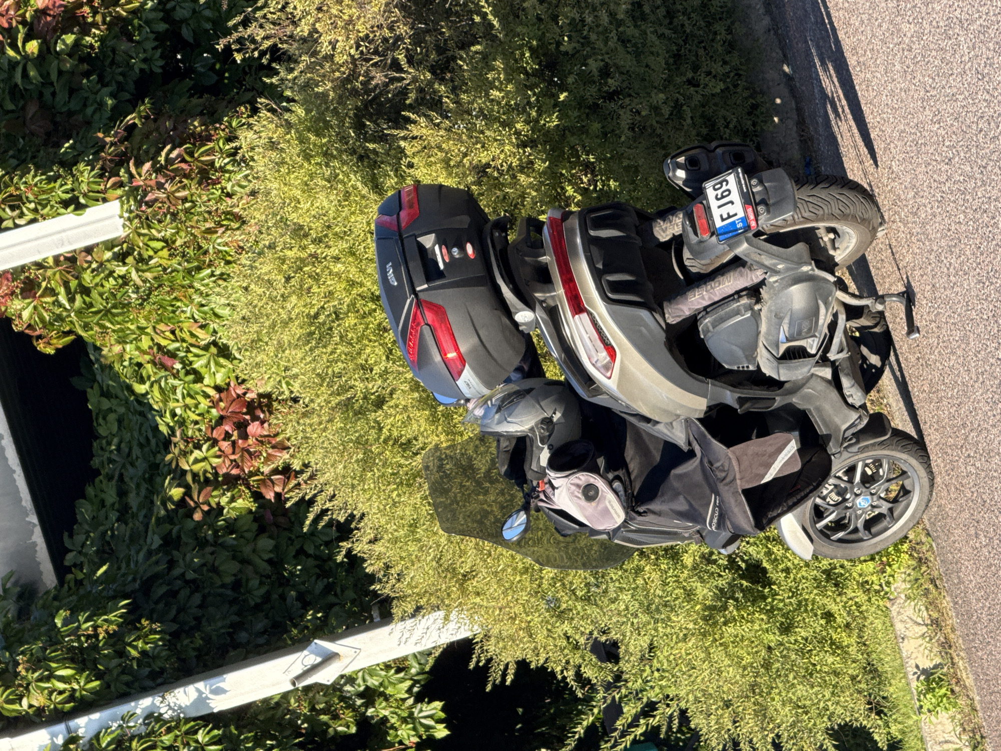 The image shows a parked three-wheeled scooter with a black top case and a silver body. The scooter is parked on the side of a road, with lush green foliage and trees in the background. A white post is visible to the left of the scooter. The license plate on the scooter reads "FJ69 ST". The scooter is facing away from the camera, and its rear wheel is on the right side of the frame. The image is taken in daylight, with shadows cast on the road, suggesting a sunny day. The location is identified as Tallinn, Estonia.
