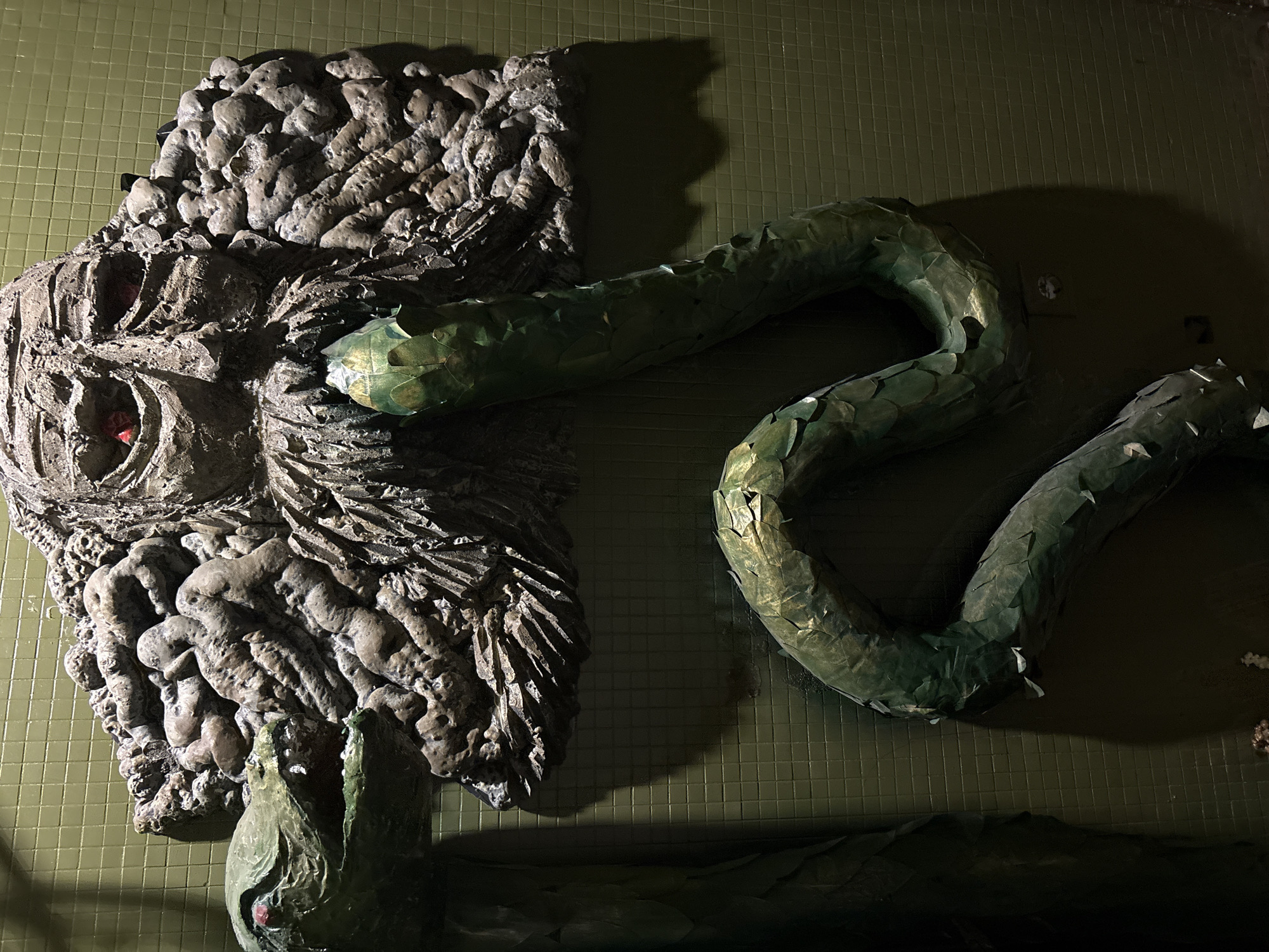 The image depicts a wall-mounted art installation. On the left is a gray, roughly textured sculpture of a monster-like face with what appears to be red coloration in the eye sockets. To the right, a green snake sculpture with scale-like details winds across the wall, with another section of the snake visible at the bottom. The installation is mounted on a wall covered in small, square, green tiles. The lighting suggests a controlled indoor environment, with strong shadows cast by the sculptures. This image is found at the Kazan Kremlin in Kazan, Russia.