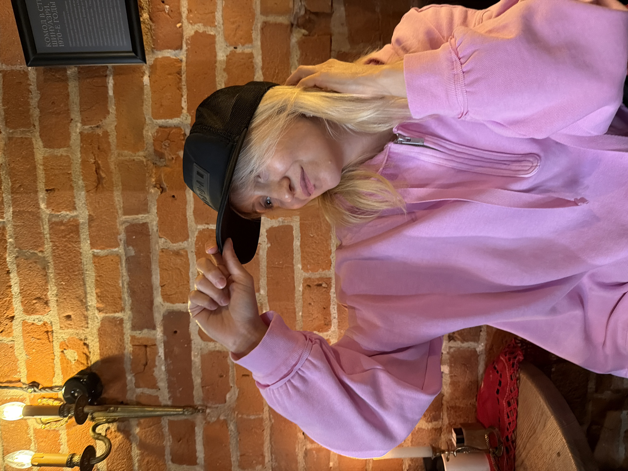 A woman wearing a black baseball cap and a pink hooded sweatshirt stands in front of a brick wall. The wall has a black framed sign and a sconce with two lightbulbs. The woman's left hand holds the brim of her cap. Her other hand is near her head. A wooden table is also visible. This image appears to be shot indoors, but specific details of the location in Moscow are not discernible from the provided image.
