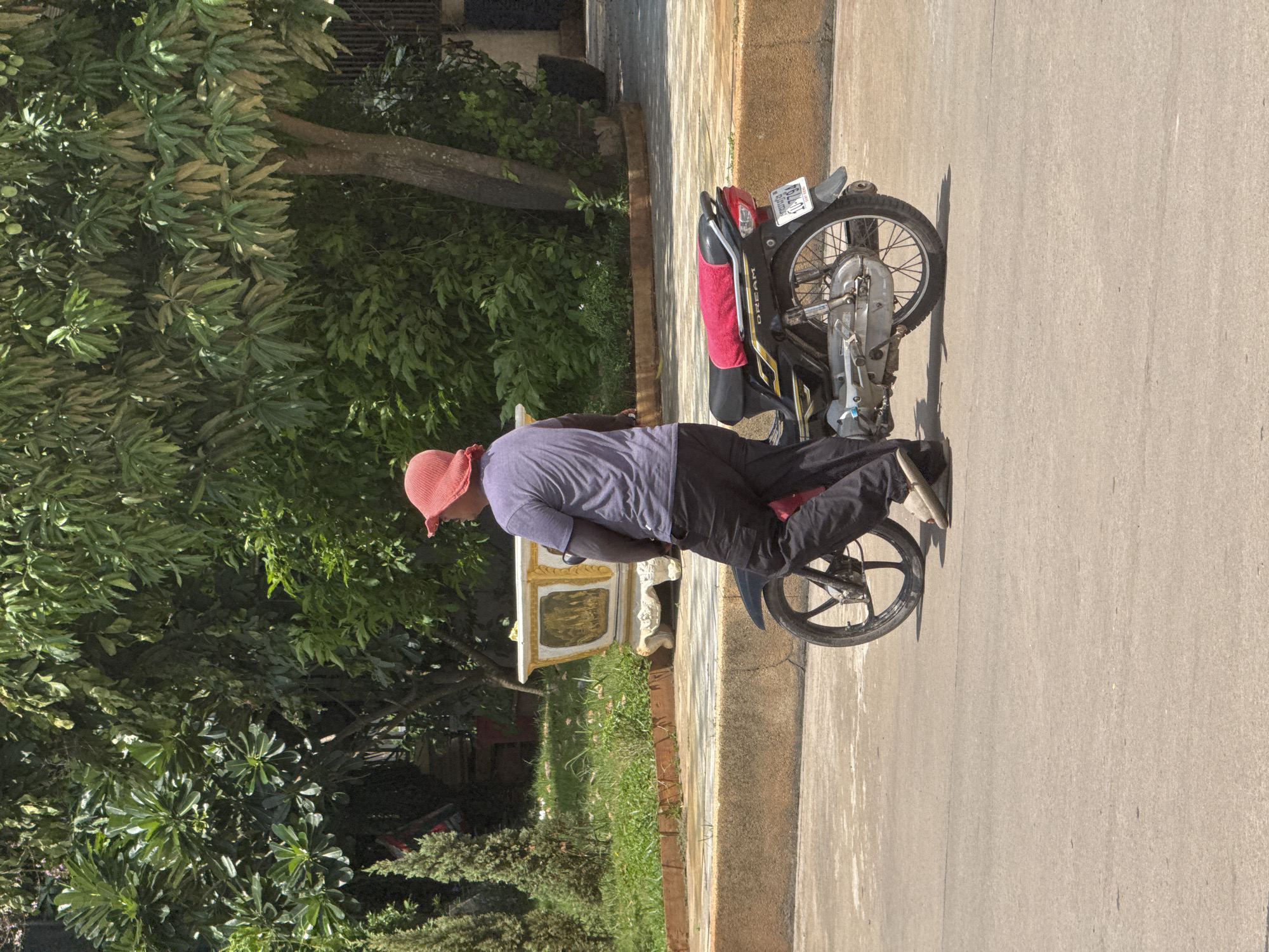 A person wearing a purple t-shirt and black pants, with a red hat, is standing next to a black motorcycle with a red towel on the seat. The motorcycle has a license plate that reads "10-7794". The scene appears to be on a paved street in front of a low wall with a grassy area behind it. Lush green trees and vegetation are visible in the background, with a decorative planter to the left of the person. The lighting suggests it is daytime and sunny. The location is identified as Bavet Quarter, Cambodia.
