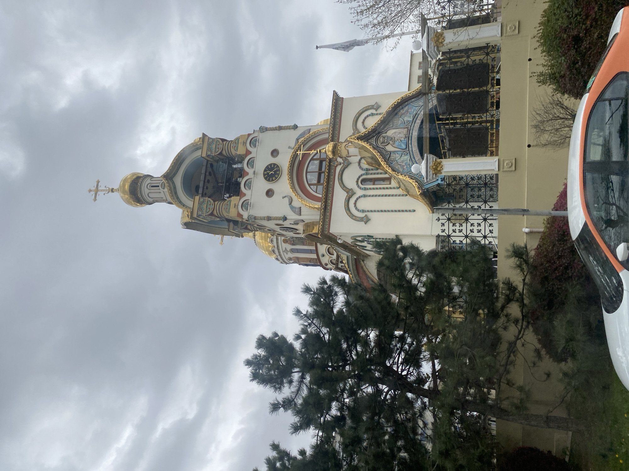 The image depicts the Church of the Holy Apostle Thaddeus in the city of Sochi, Russia. The church has a traditional Orthodox architectural style, featuring a golden dome topped with a cross. A clock tower is visible on the church's facade. The church's exterior is decorated with ornate patterns, including gold trim, along the roofs and the windows. The trees in front of it fill most of the frame. The sky is overcast with heavy clouds.