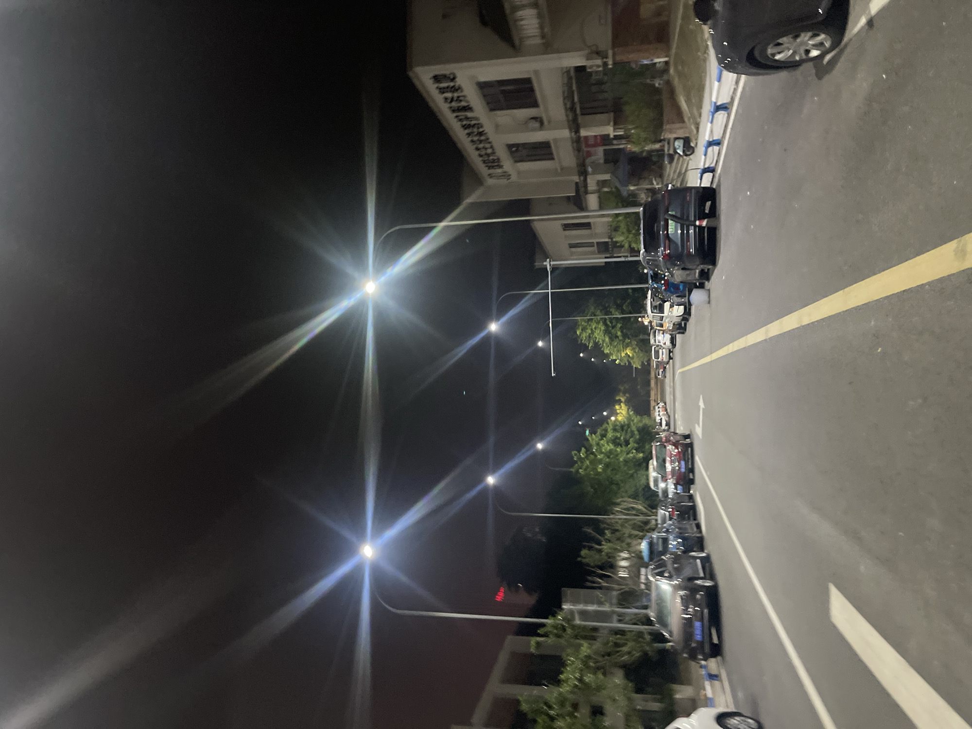 The image depicts a street scene at night in the Chongqing High-tech Zone, China. The street is illuminated by streetlights, which create bright flares in the image. Several cars are parked along the side of the road. Buildings are visible on the right side of the street, one of which has Chinese characters on its facade. Trees and other vegetation line the left side of the street. The overall scene is quiet and appears to be a typical urban environment at night.