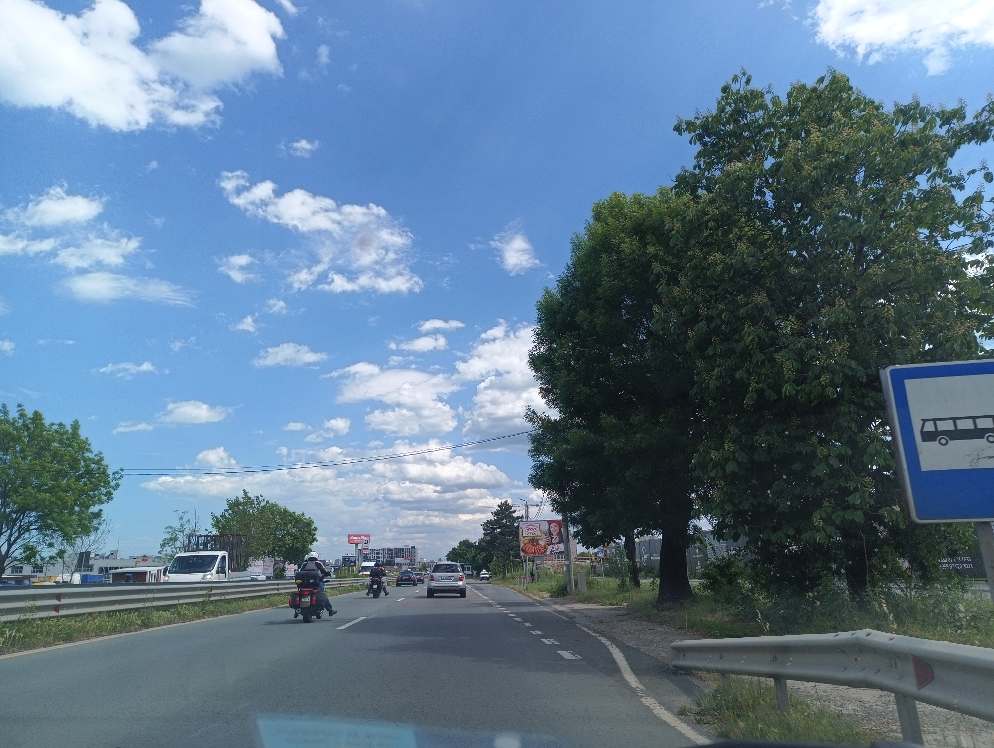 The image depicts a road in Burgas, Bulgaria, under a blue sky with scattered clouds. The road has two lanes with a broken white line separating them. A car and two motorcycles are traveling in the same direction, away from the camera. On the right side of the road, there are trees and a bus stop sign. On the left, there's a roadside barrier and greenery, with buildings and billboards visible further in the distance.