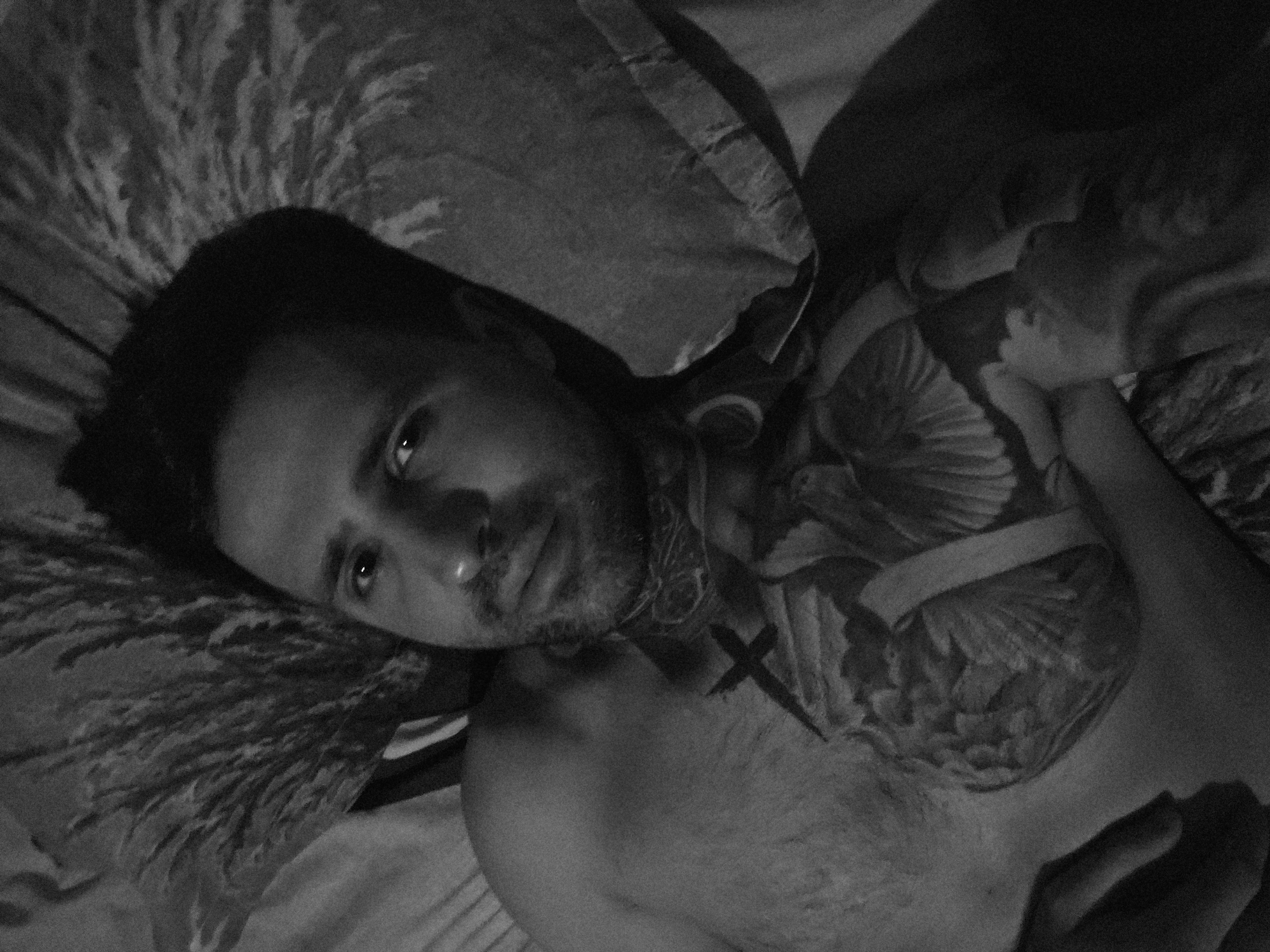 A black and white, high-angle, close-up shot shows a person lying on their back in bed. The person is a male with short, dark hair and a goatee. He is shirtless, revealing a large tattoo of a heart with wings on his chest. He is wearing a patterned bandana around his neck. The background consists of a pillow with a floral or feather pattern. The lighting is dim, creating shadows and highlighting the textures of the bed linens and the tattoo. There is no discernible indication of the specific location within Stara Zagora, Bulgaria.