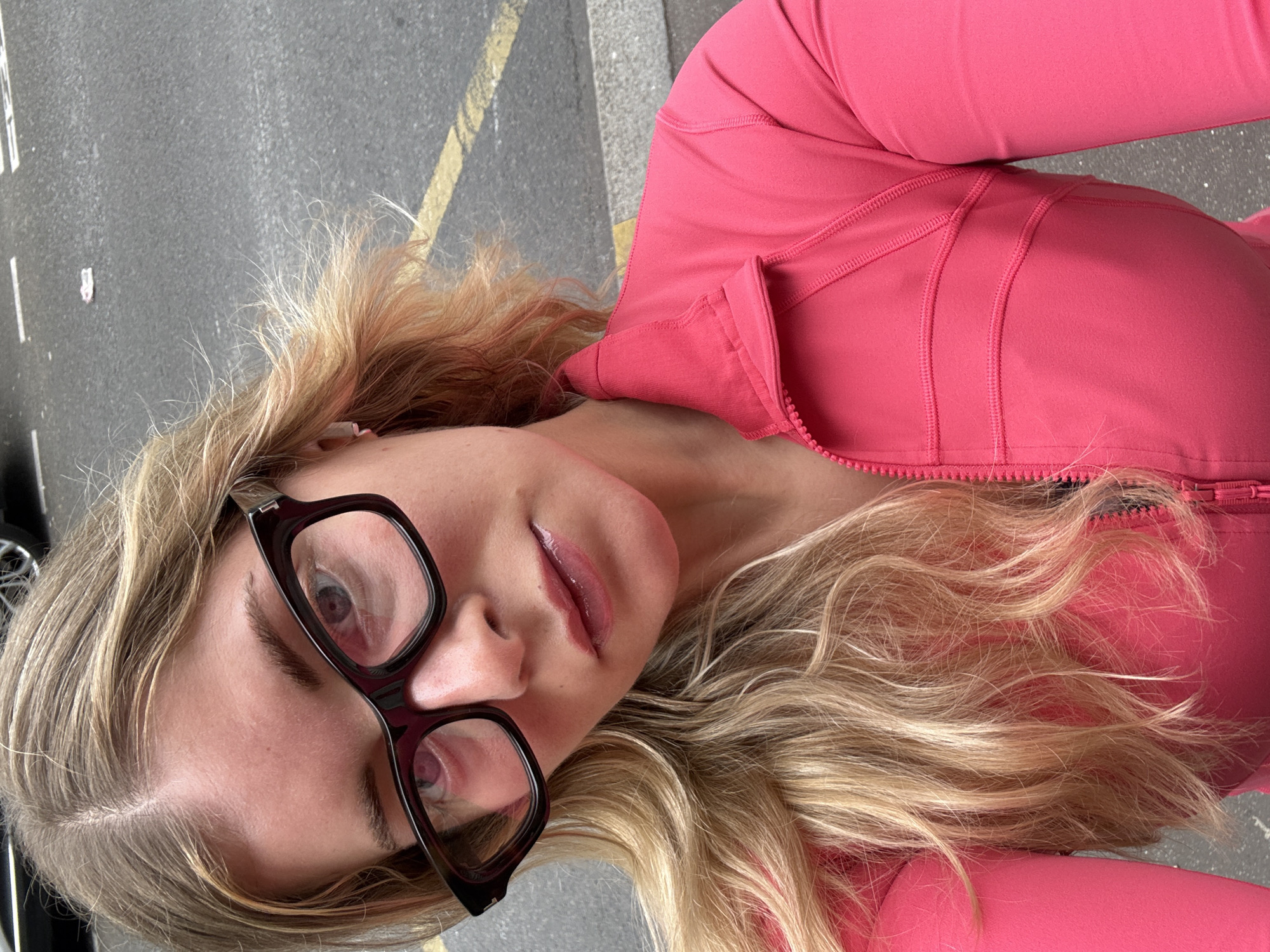 A woman with blonde hair and black-framed glasses is seen in a close-up, slightly angled shot. She is wearing a pink zip-up jacket. The background appears to be a street in Paris, France, with visible asphalt and yellow lane markings.