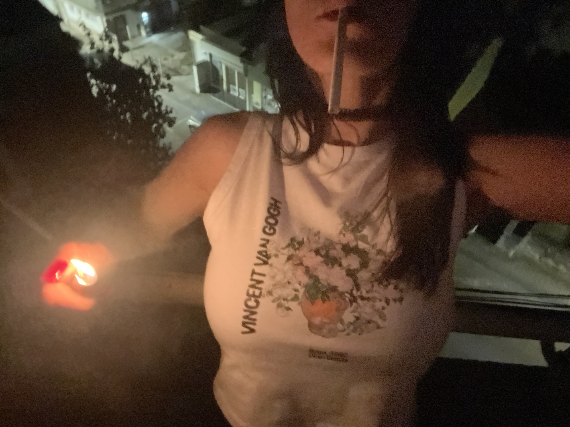 A low-light, eye-level photograph captures a person, likely female, from the chest up, at night. The subject appears to be outdoors, possibly on a balcony or terrace, overlooking dimly lit buildings and dark foliage in the background, indicative of an urban or suburban setting.
The person is wearing a white sleeveless top. Prominently displayed on the left side of the shirt, vertically aligned, is the text "VINCENT VAN GOGH". To the right of this text, a colorful print of a floral arrangement in a vase is visible. Faintly visible below the floral design are additional words, possibly "FLOWER POT" and the year "1880". The person also wears a dark choker necklace.
In their left hand, a red lighter is actively aflame, casting a warm glow on their hand and arm. A slender, light-colored object, consistent with a cigarette or similar item, is held in their mouth or near their nose, suggesting the act of lighting it. The overall scene is dark, with the lighter's flame providing the primary illumination for the foreground activity, while distant artificial lights can be seen from the background buildings.
