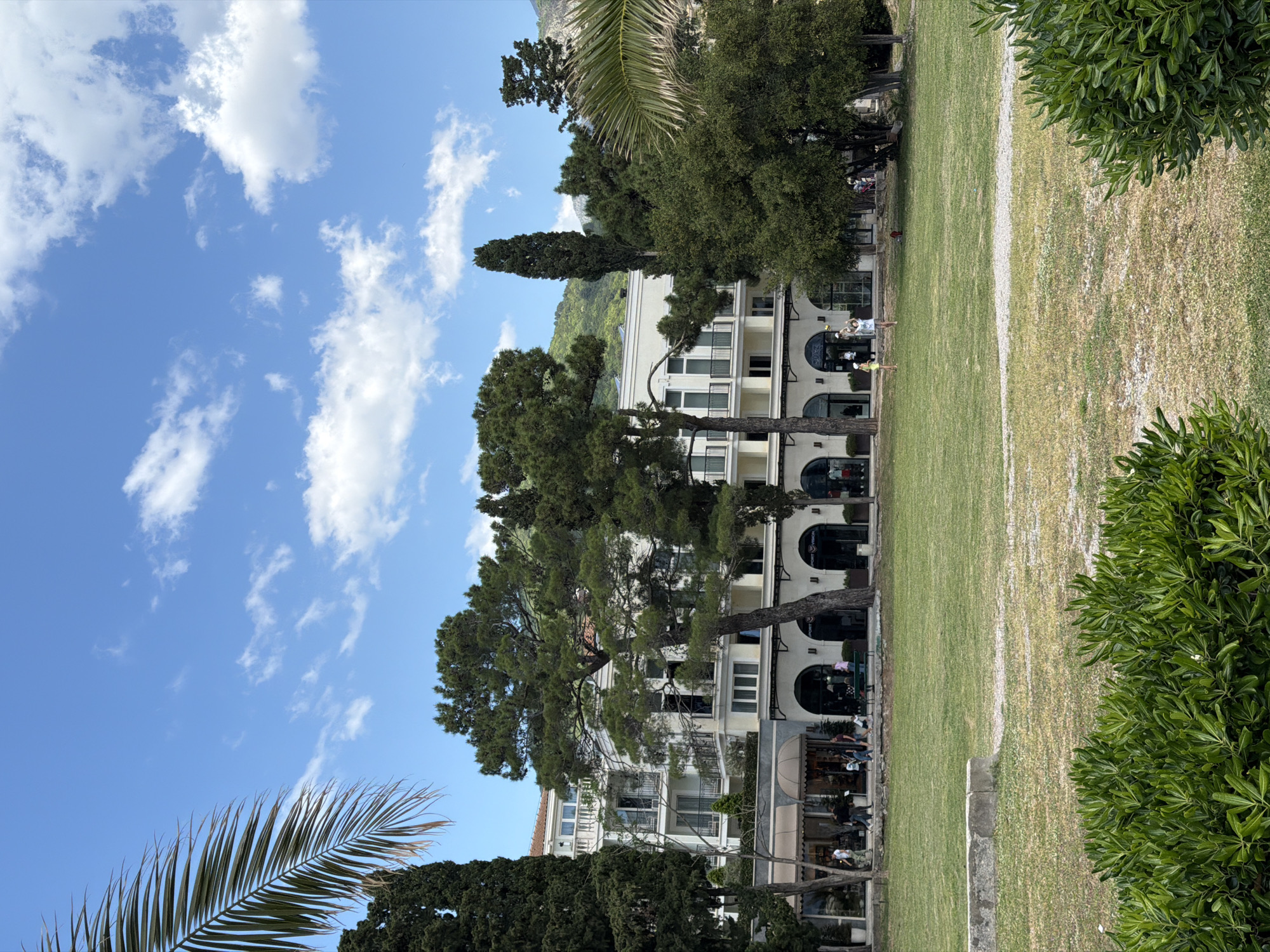 The image shows a partial view of a multi-story building with arched windows and white façade, obscured by tall pine trees. The building faces a long, green lawn divided by a white line. People are visible on the building's ground floor, possibly sitting at tables. Palm and cypress trees frame the scene. The sky is blue with scattered white clouds. The image depicts a scene in Budva, Montenegro.