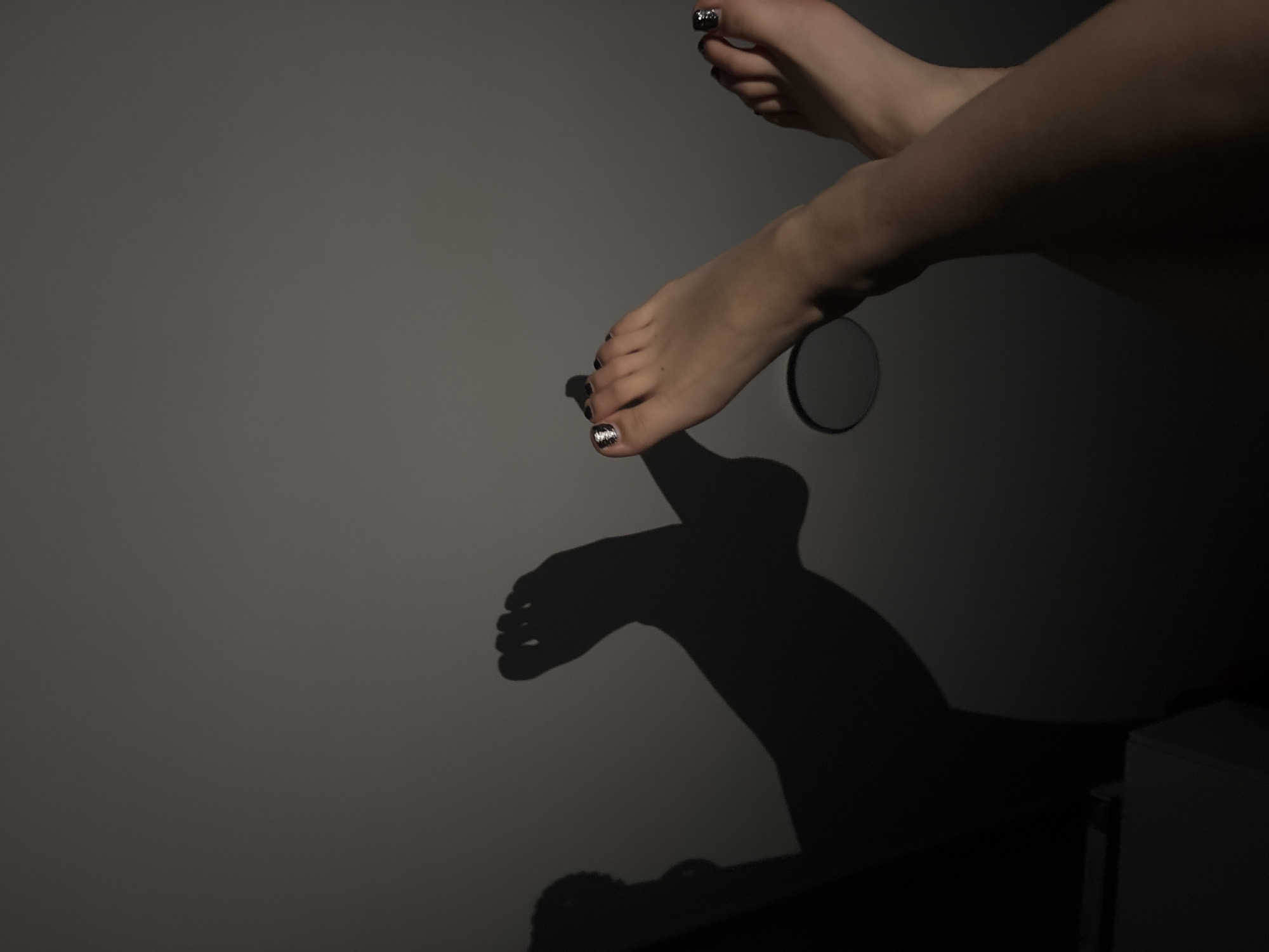 A dimly lit photograph shows two human feet resting on a wall. The feet have decorated toenails. A small, round mirror is placed next to one of the feet. Shadows of the feet are cast onto the wall. A corner of a cabinet is visible in the lower right corner. The location is unknown.