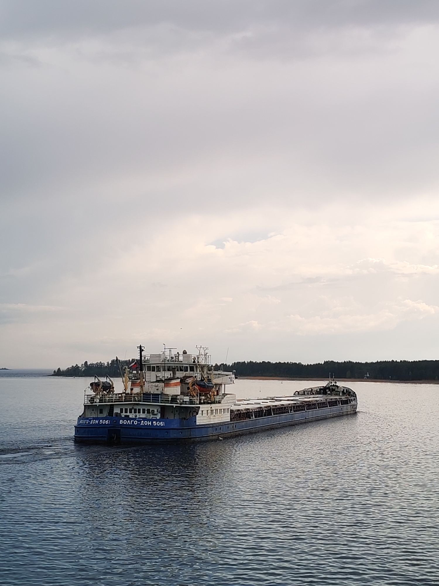 A long cargo barge labeled "Volgo-Don 5061" is traversing a large body of water. The barge is blue and white and has a multi-tiered structure at the stern. The water is dark blue and ripples slightly from the vessel's passage. The sky is overcast, casting a soft light across the scene. In the distance, a shoreline with trees is visible. The barge is traveling in the area of the city of Shalsky, Russia.