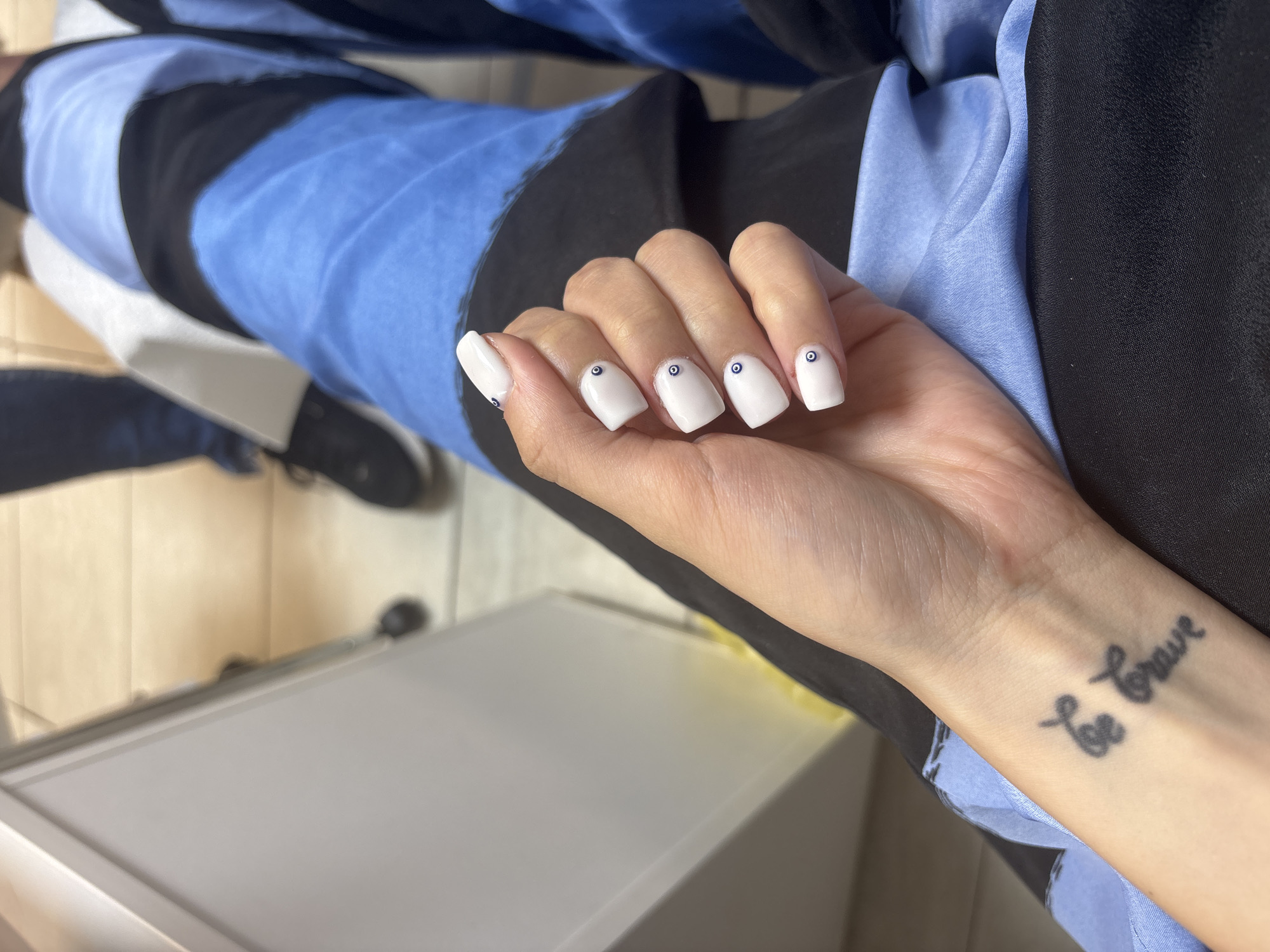 The image depicts a close-up of a person's hand with white square-shaped nails, each adorned with a small blue and black eye design. The hand is positioned against a backdrop of clothing featuring a pattern with shades of blue and black. The person has a "Be brave" tattoo on their wrist. A portion of the person's legs, clad in the same patterned clothing, is visible in the background, along with a glimpse of a black shoe. In the background, elements of an interior space are visible, including a white cabinet and flooring. The exact location within the city of Sofia, Bulgaria, is not ascertainable from the provided image.