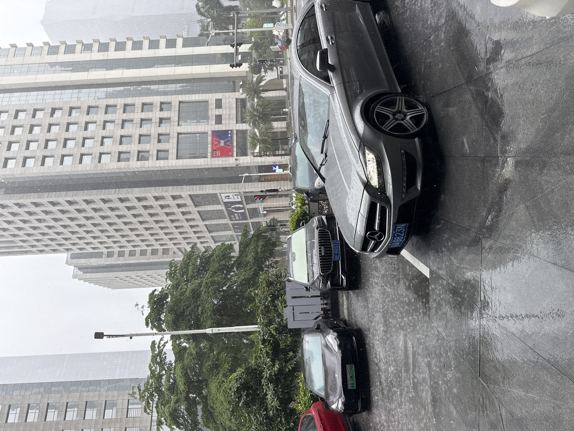 This image depicts a street scene in Tianhe District, China, during heavy rainfall. Multiple vehicles are parked on a wet, tiled surface, with reflections visible on the ground. In the foreground, a silver Mercedes-Benz sedan is prominent. To its left, a black Tesla sedan is parked, alongside a dark-colored SUV. Further left, a red car is partially visible.
Behind the vehicles, lush green trees and foliage line the sidewalk. Towering modern buildings with multiple windows dominate the background, characteristic of an urban environment. Traffic lights and street lamps are also present. The sky is overcast, contributing to the gloomy atmosphere, and the falling rain is clearly visible as streaks against the buildings and vehicles. No people are clearly discernible in the scene. The overall impression is of a busy city street experiencing inclement weather.