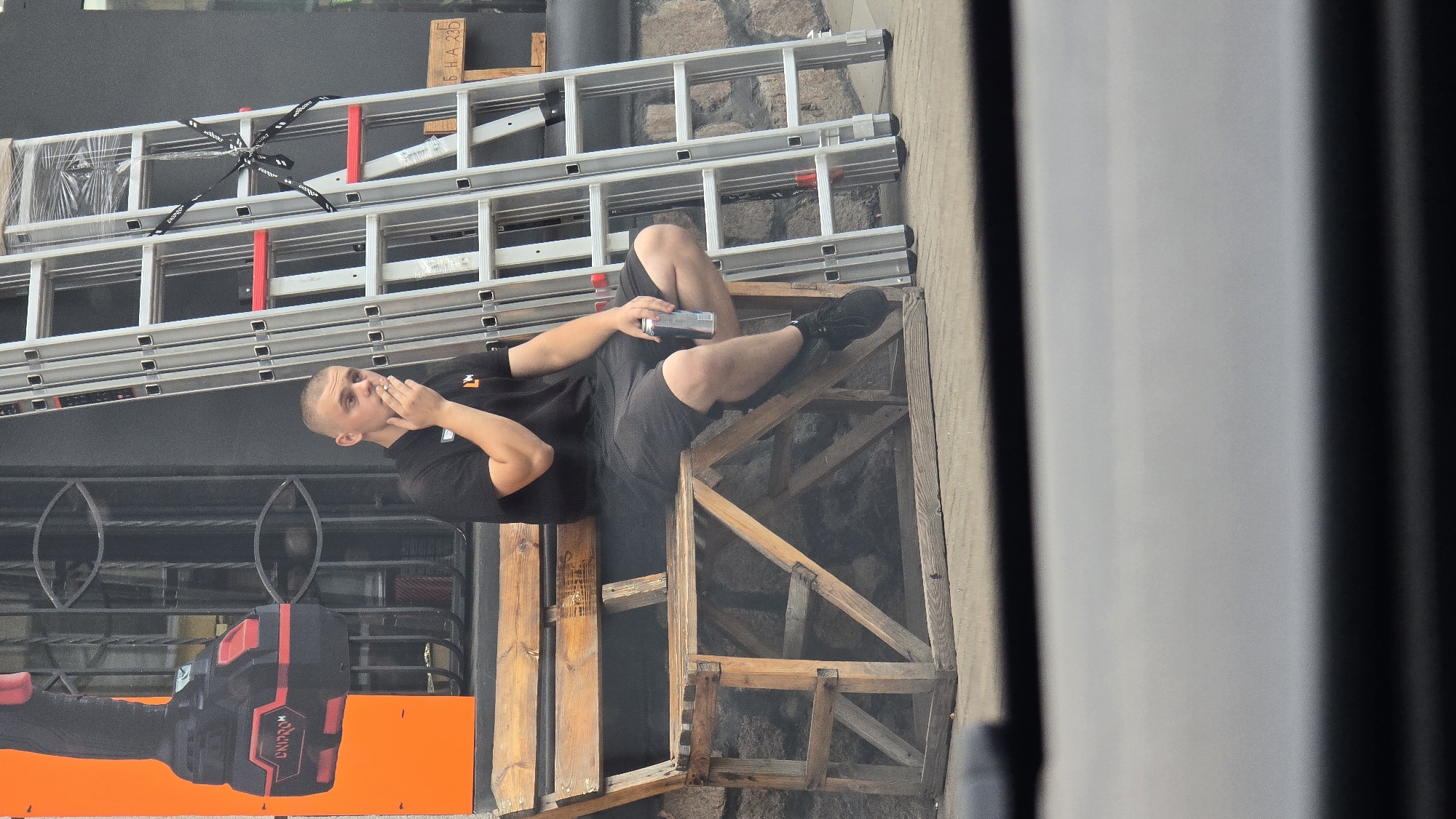 A young man is sitting on a wooden bench outside a building, smoking a cigarette and holding a can. He is wearing a black t-shirt and black shorts, with black sneakers. Behind him, a tall silver ladder is leaning against the building. To the left of the man, a large orange advertisement featuring a power tool is displayed on the building's facade. The scene appears to be set on a street in Zhytomyr, Ukraine.