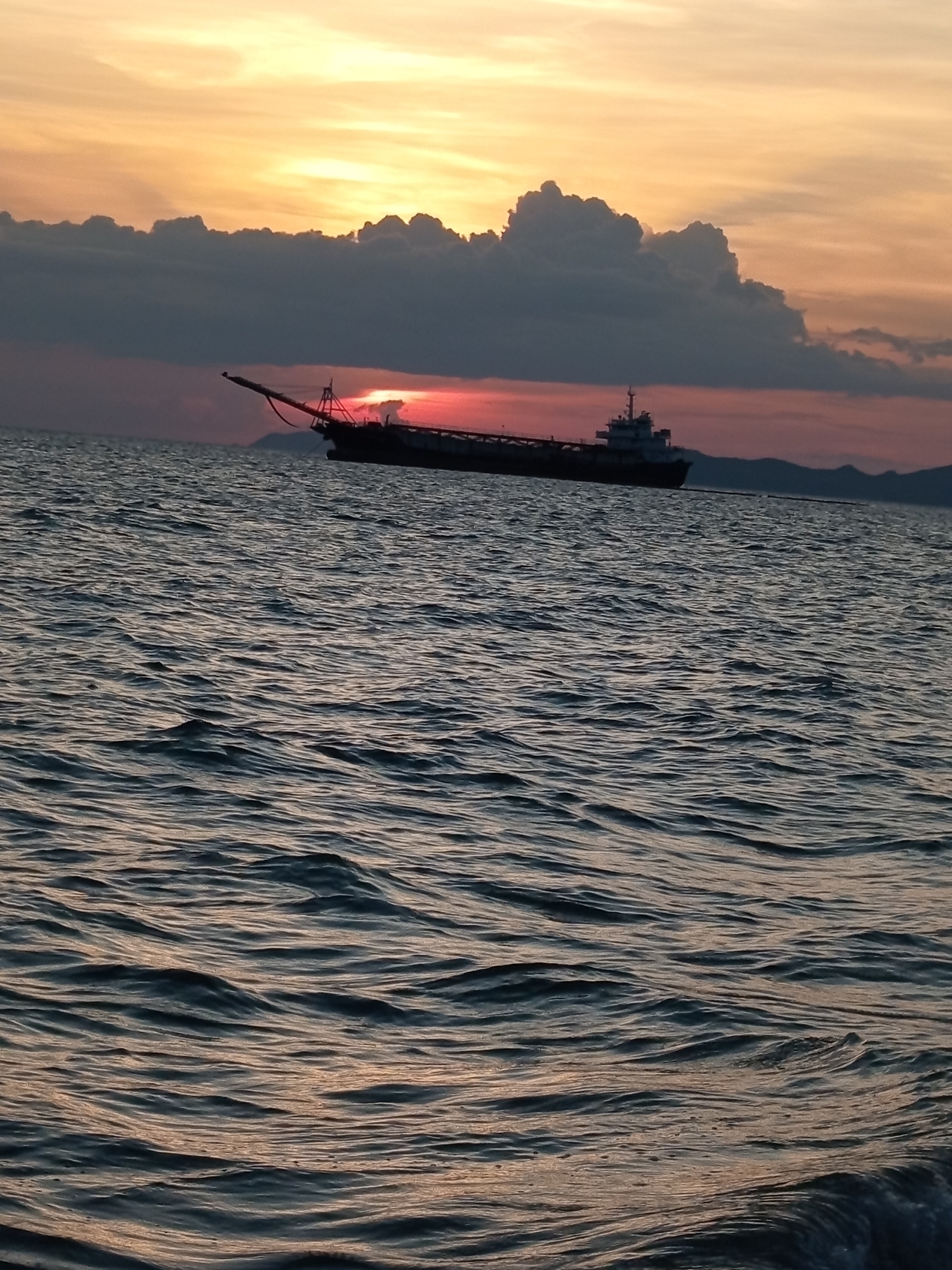 A cargo ship is silhouetted against a sunset over the ocean in Pattaya City, Thailand. The sky is a mix of orange, yellow, and pink hues, with dark clouds in the upper portion. The ship is positioned in the middle ground, with its stern facing left and its bow extending towards the center of the frame. Waves ripple across the dark water in the foreground. The scene is serene and depicts the natural beauty of a coastal sunset.