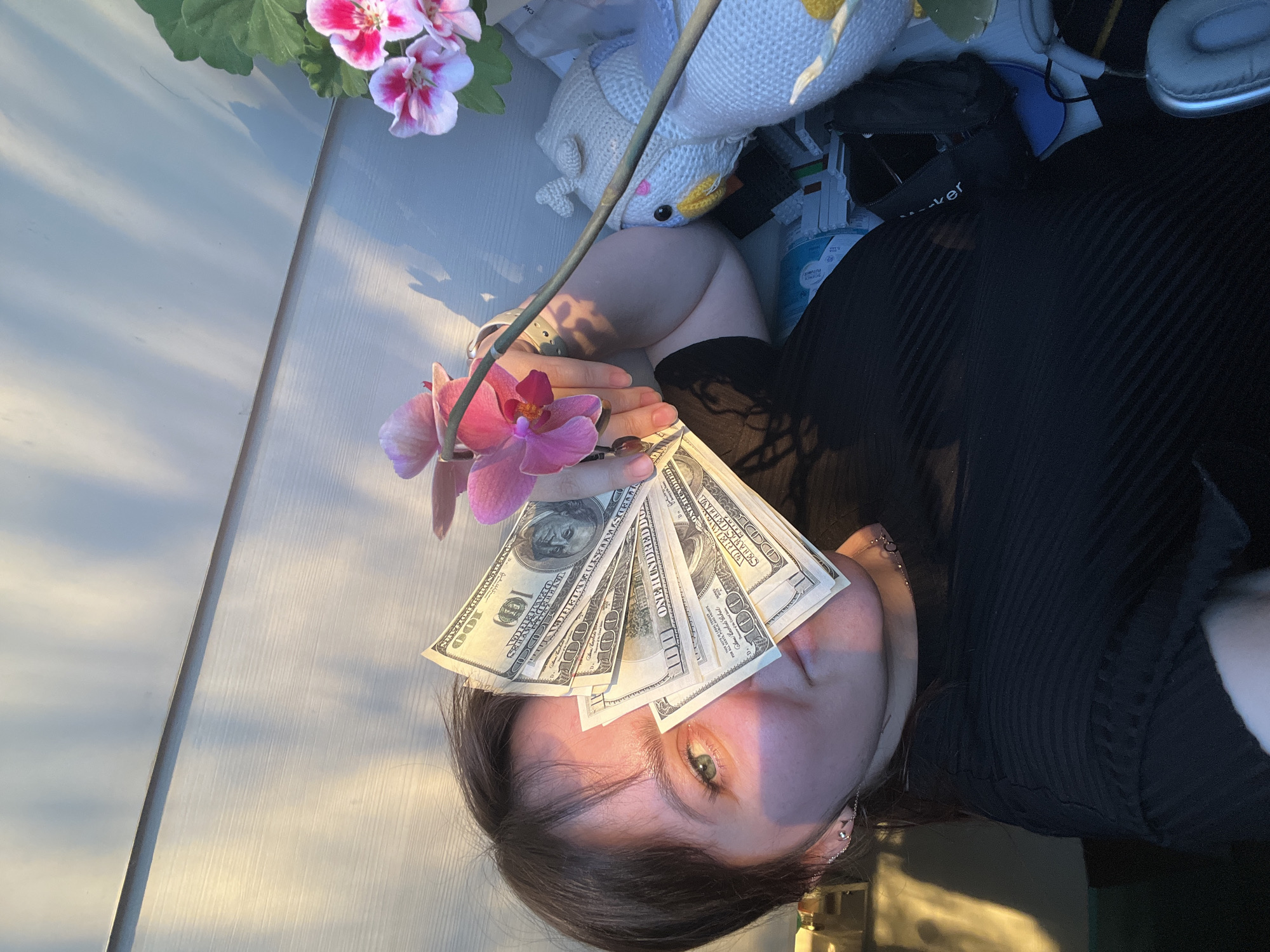 A woman lies on a table holding a bunch of one-hundred-dollar bills and an orchid, positioned on her forehead and over her face. The table appears to be indoors. Other objects around her include a stuffed animal, a cleaning supply container, headphones, and pink flowers in a pot. Her right hand is holding the stem of the orchid. She is wearing a black shirt. The location is not discernible as being in the city of Noviy, Russia from the visible evidence.