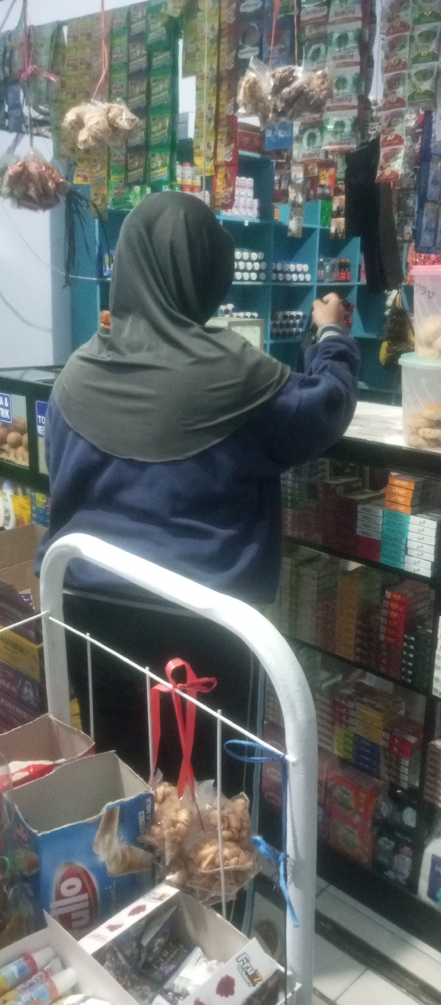 A person wearing a dark gray hijab and a dark blue jacket is standing in front of shelves stocked with various goods in a small store. The store appears to be in Kediri City, Indonesia, as indicated by the context. The shelves are filled with packaged snacks, drinks, and other retail items. In the foreground, there is a white rack with some boxes and bags of goods. One bag contains light brown, irregularly shaped snacks. A red ribbon is tied to the rack. The overall impression is of a cluttered but functional retail environment.