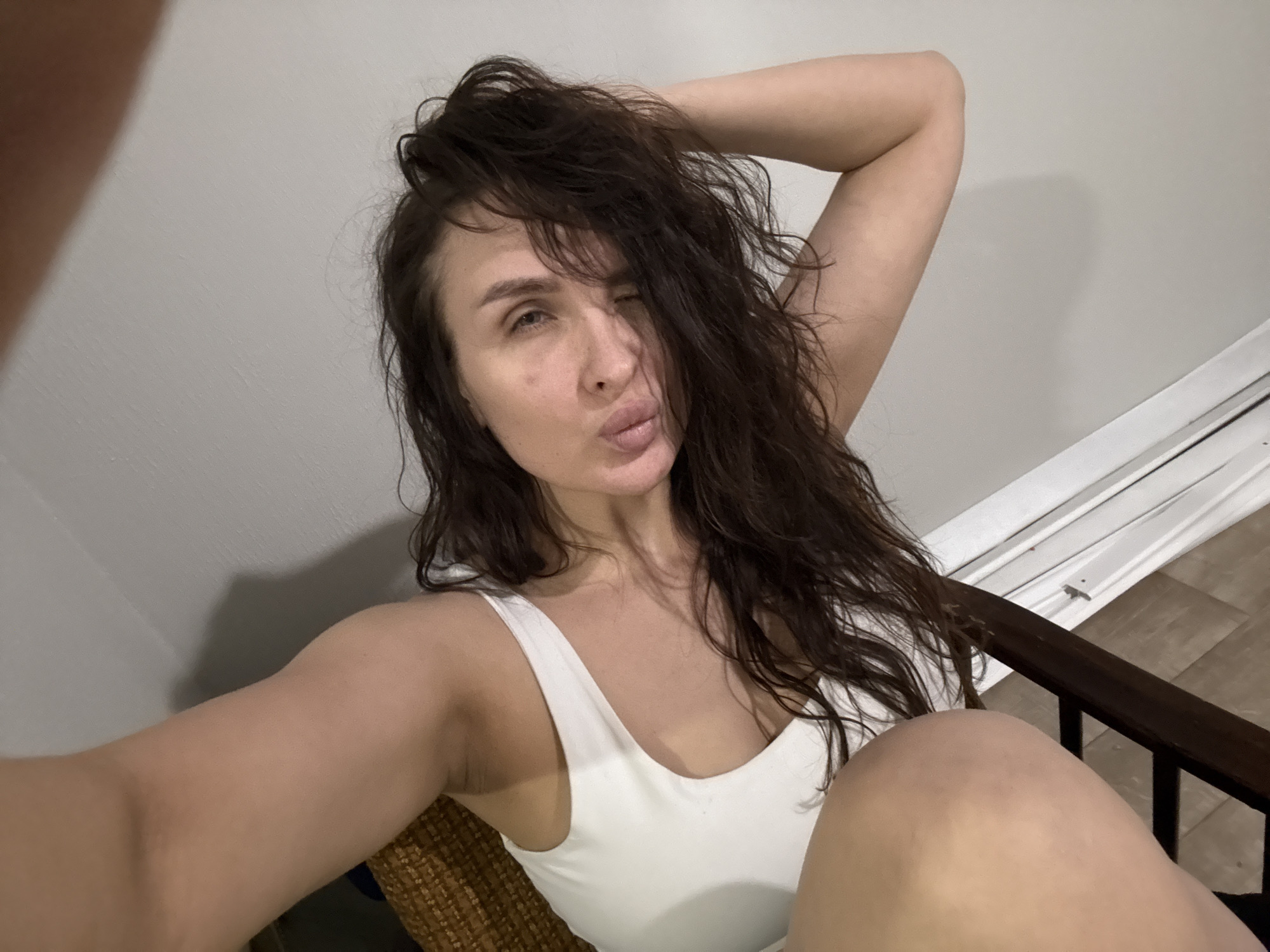 A woman with dark hair, wearing a white tank top, poses in a chair against a light gray wall. She is winking with her left eye, and her lips are pursed in a kiss. Her right arm is raised and bent, with her hand behind her head. Part of a window or door frame is visible in the background to the right. The location is not discernible as Chaikovsky, Russia, from the provided image.