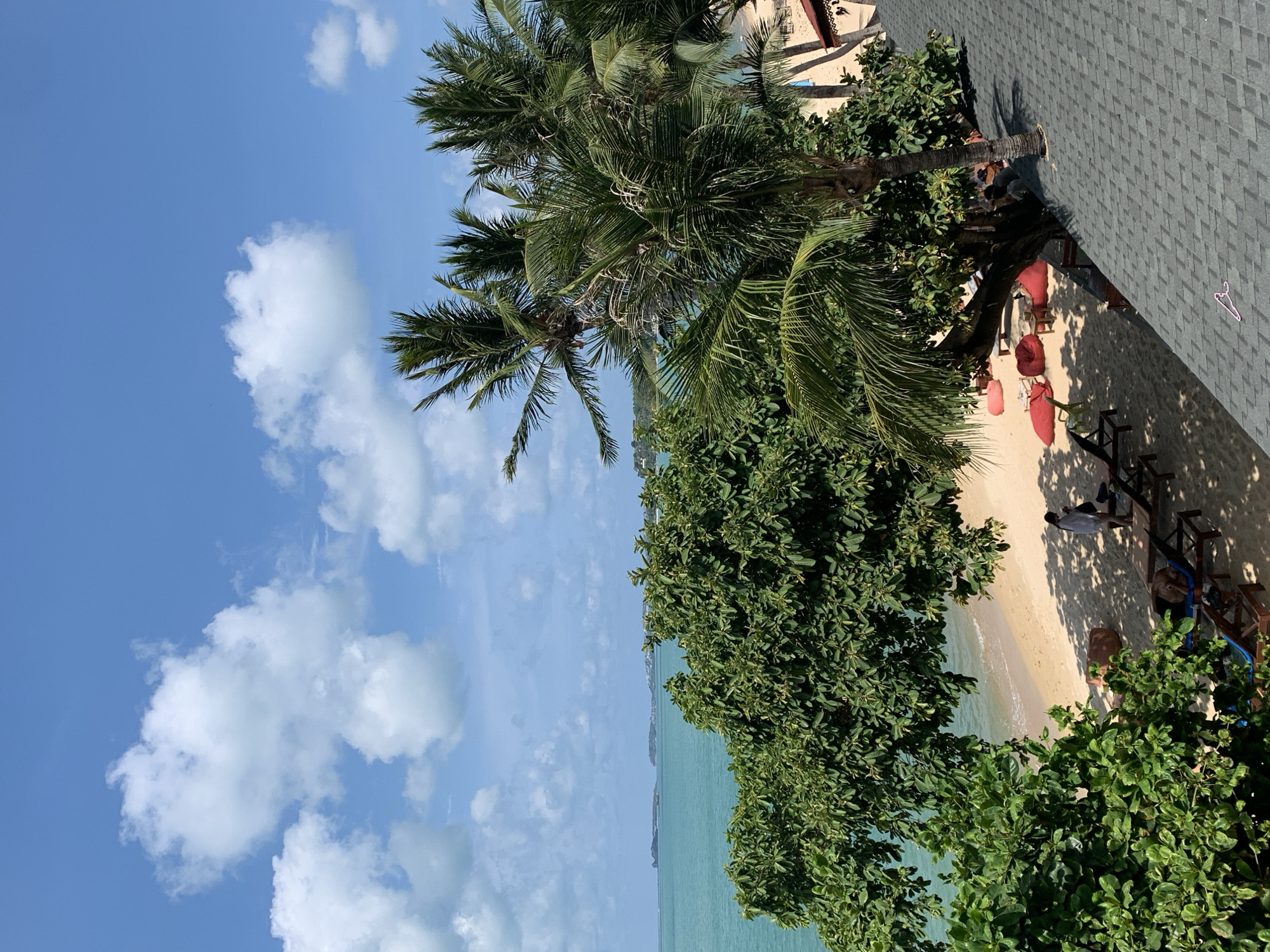 The image captures a vibrant, sunny tropical beach scene, viewed from an elevated perspective, likely a balcony or rooftop. The setting is Baan Mae Nam, Thailand, characterized by its pristine sandy beach, calm turquoise ocean, and lush green tropical vegetation, including several prominent palm trees.
Under a bright blue sky adorned with fluffy white cumulus clouds, the beach shows signs of leisure and relaxation. Several red beanbag chairs and wooden lounge chairs, some with blue cushions, are scattered on the sand, predominantly within the shade cast by large trees. A few individuals are visible on the beach; one person in a light-colored shirt stands near a lounge chair, while another appears to be relaxing on a chair. Further down the beach, other faint figures suggest more activity under the trees.
The photo was taken looking down and across the beach. In the foreground on the right, a dark, shingled rooftop is visible, with a distinctive pink clothes hanger-shaped object resting on it. The overall impression is one of a peaceful and inviting tropical getaway, indicating a pleasant mid-day with perfect weather for beach activities. No visible text is present in the image.