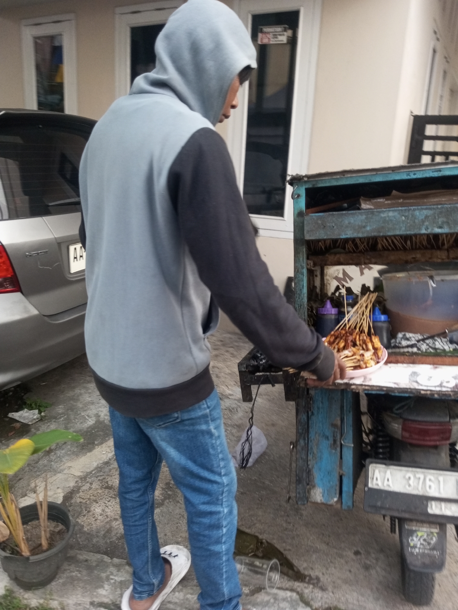 In Pandansari, Indonesia, a person wearing a gray hooded sweatshirt and blue jeans stands next to a blue food cart. The food cart appears to be a mobile vendor, possibly selling satay, as skewers of meat are visible on a plate within the cart. The cart is attached to the rear of a motorcycle, indicated by the visible tire and license plate reading "AA 3761". A silver car with a license plate "AA" is parked to the left of the scene. A potted plant with large green leaves is situated on the ground to the left of the food cart. The ground is paved with concrete. The background features a building with white window frames.