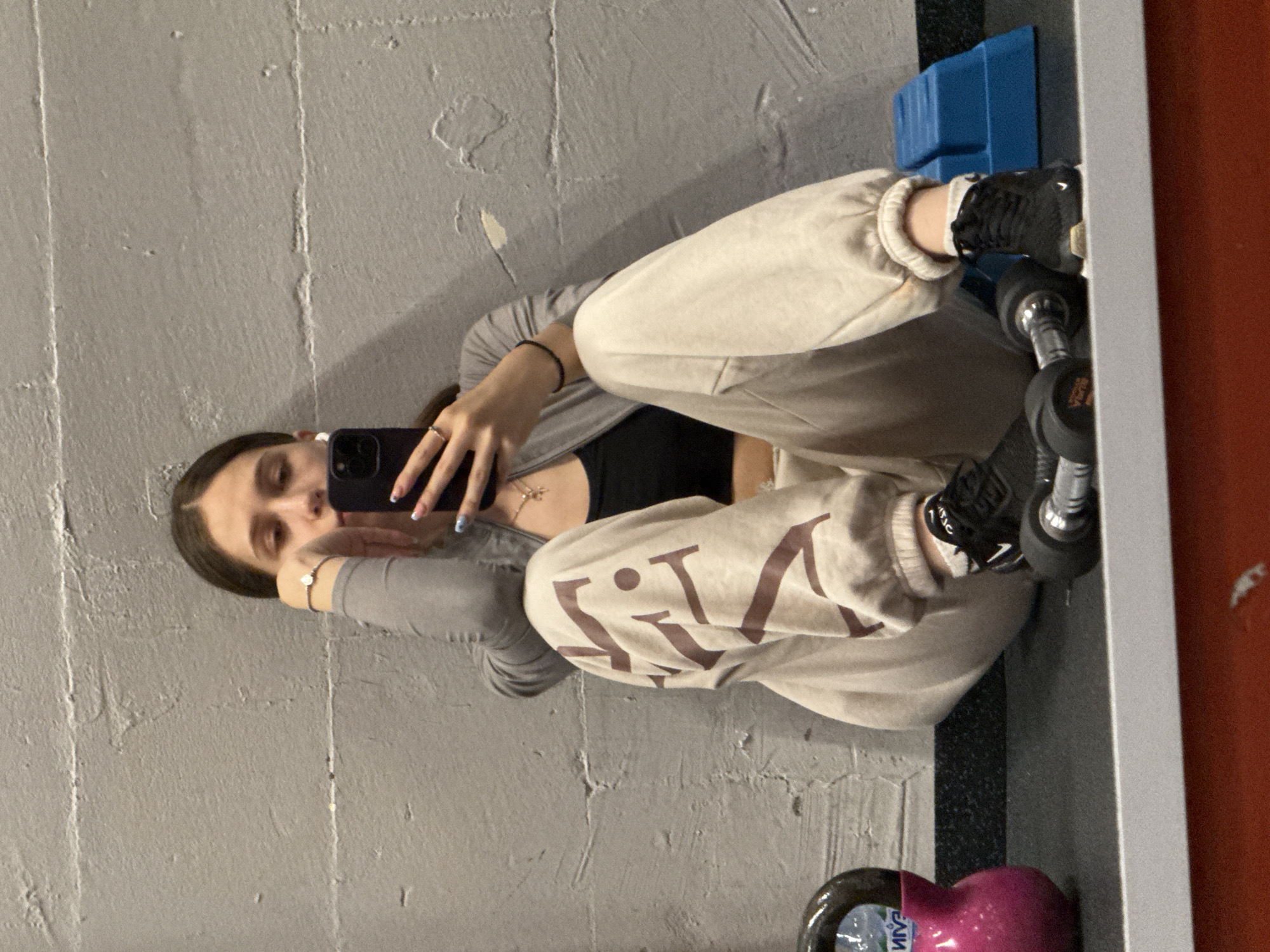 An indoor scene in Sofia, Bulgaria, depicts a young female sitting on a dark grey floor, leaning against a textured light grey concrete wall. She is wearing a grey long-sleeved top over a black sports bra, light beige sweatpants with visible brown "VIT" lettering on the left leg, white socks, and black athletic shoes with a white Nike swoosh on the right shoe. Her left wrist displays a silver bracelet with a central clear stone, and her right ring finger wears a silver ring. A gold-colored cross pendant hangs around her neck. Her nails are painted light blue with white tips. She holds a dark-colored smartphone, likely an iPhone with triple cameras, in her right hand, angled as if taking a mirror selfie. Her left hand rests on her temple in a contemplative pose, and her gaze is directed towards the phone screen.
On the left side of the frame, a pink kettlebell with a partially visible, inverted label "ИЙVЭ" (suggesting a brand like "DEVIN") is present. Directly in front of her feet, two dumbbells with black 1 kg plates and silver handles are resting on the floor. A blue aerobic step platform is positioned behind her right foot. The bottom edge of the image is defined by a reflective surface, possibly a mirror or polished metal, showcasing partial reflections of the floor and equipment. The concrete wall exhibits textural variations, horizontal lines, and areas of minor paint peeling or discoloration. The lighting is diffused and even, illuminating the scene without harsh shadows.
